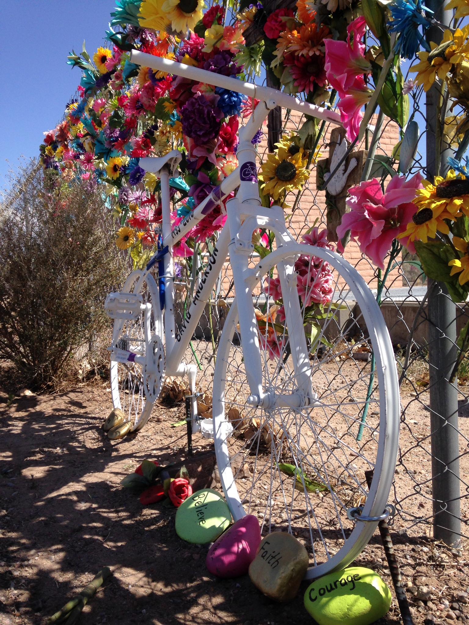 A white bicycle surrounded by a colorful array of artificial flowers on a fence, with painted rocks labeled "Believe," "Faith," and "Courage" placed at the base of the bike, all set against a clear blue sky. Albuquerque Bosque mountain bike trail.