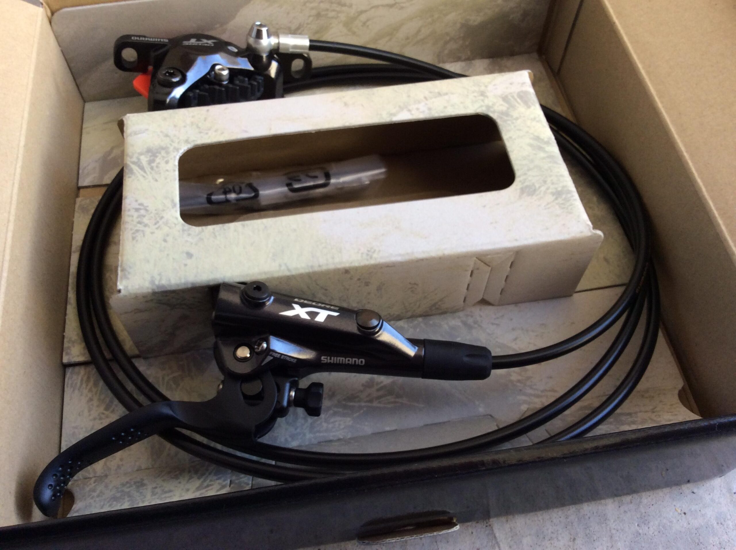 A Shimano XT bicycle hydraulic brake system, including brake lever, caliper, and connecting hoses, displayed in an open cardboard packaging.