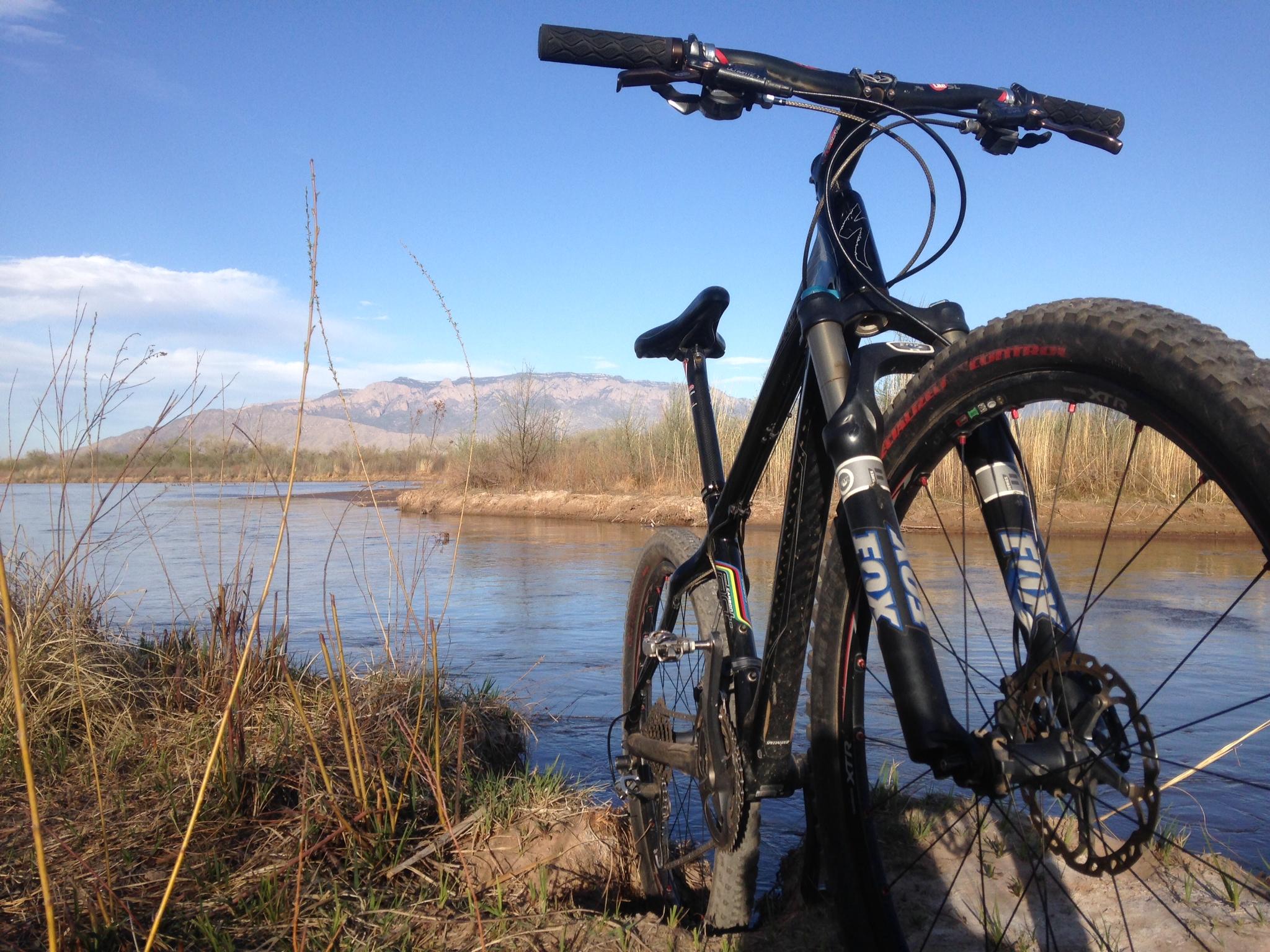 A close-up view of a mountain bike leaning against a grassy bank beside a river. In the background, there are rolling mountains under a clear blue sky. The scene captures a peaceful outdoor environment, with tall grass and the gentle ripples of the water reflecting the landscape. Albuquerque Bosque mountain bike trail.