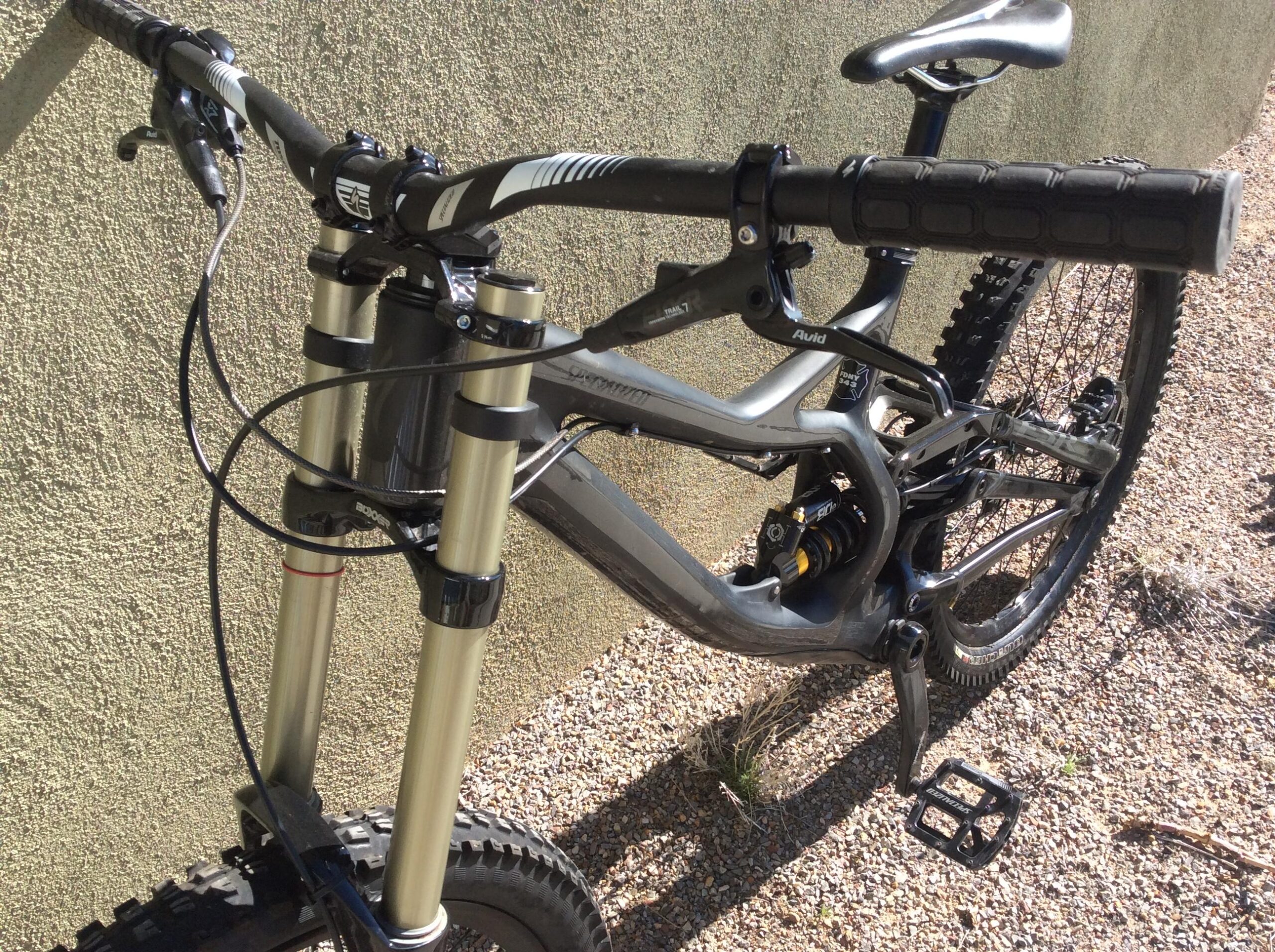 Specialized S-Works Demo 8: Mountain bike leaning against a textured wall, showcasing its handlebars, front suspension, and frame. The bike features metallic gold forks, black grips, and a detailed gear and brake system. The ground is covered with small pebbles and sparse grass.