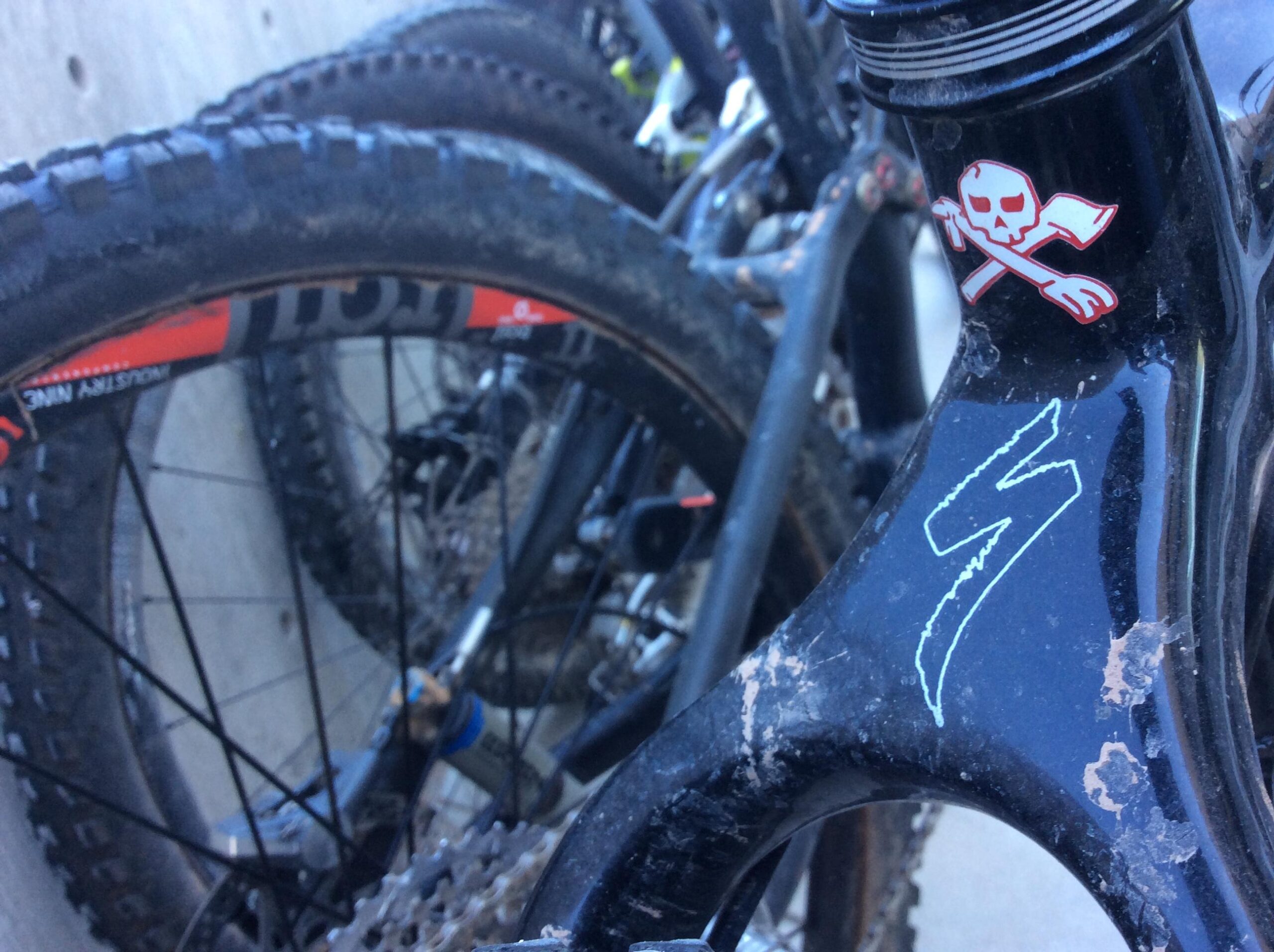 A close-up image of a mountain bike's frame and front tire, showing a black frame with a skull and crossbones sticker, markings on the frame, and the textured, muddy surface of the tire behind it. The background features additional bicycles partially obscured.