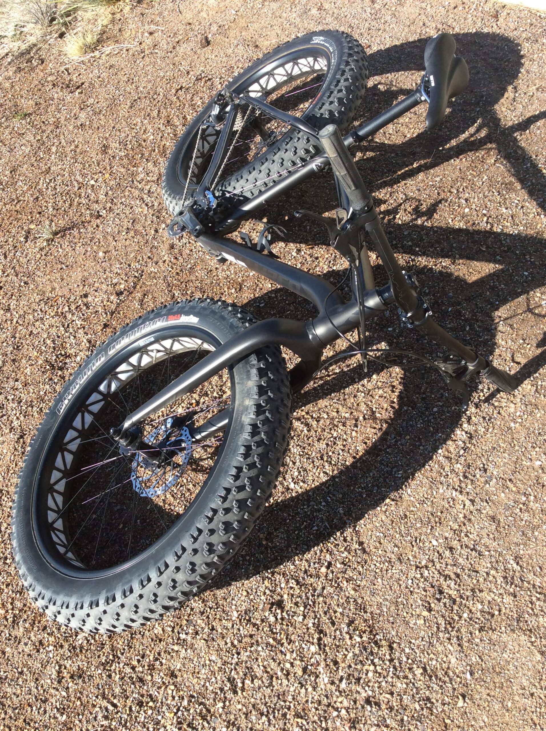 Specialized Fatboy: A black fat bike with oversized tires lying on uneven gravel terrain. The bike is positioned upside down, showing its frame, handlebars, and two large wheels. Sunlight casts distinct shadows on the ground, highlighting the bike's design features.