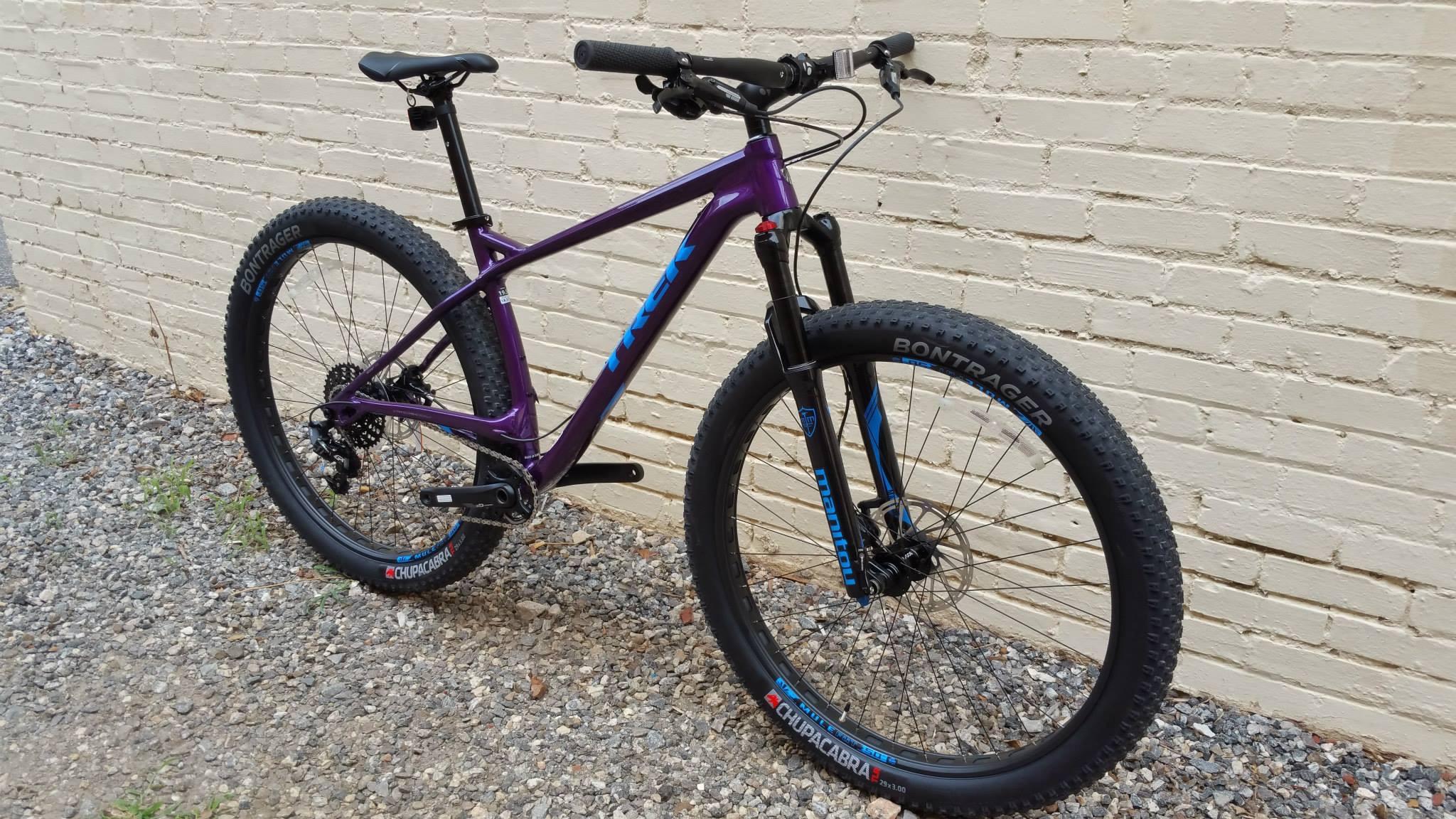 Trek Stache: A purple mountain bike with blue accents, featuring wide tires and a sturdy frame, positioned against a white brick wall. The bike has a single gear setup and a suspension fork, resting on a gravel surface.