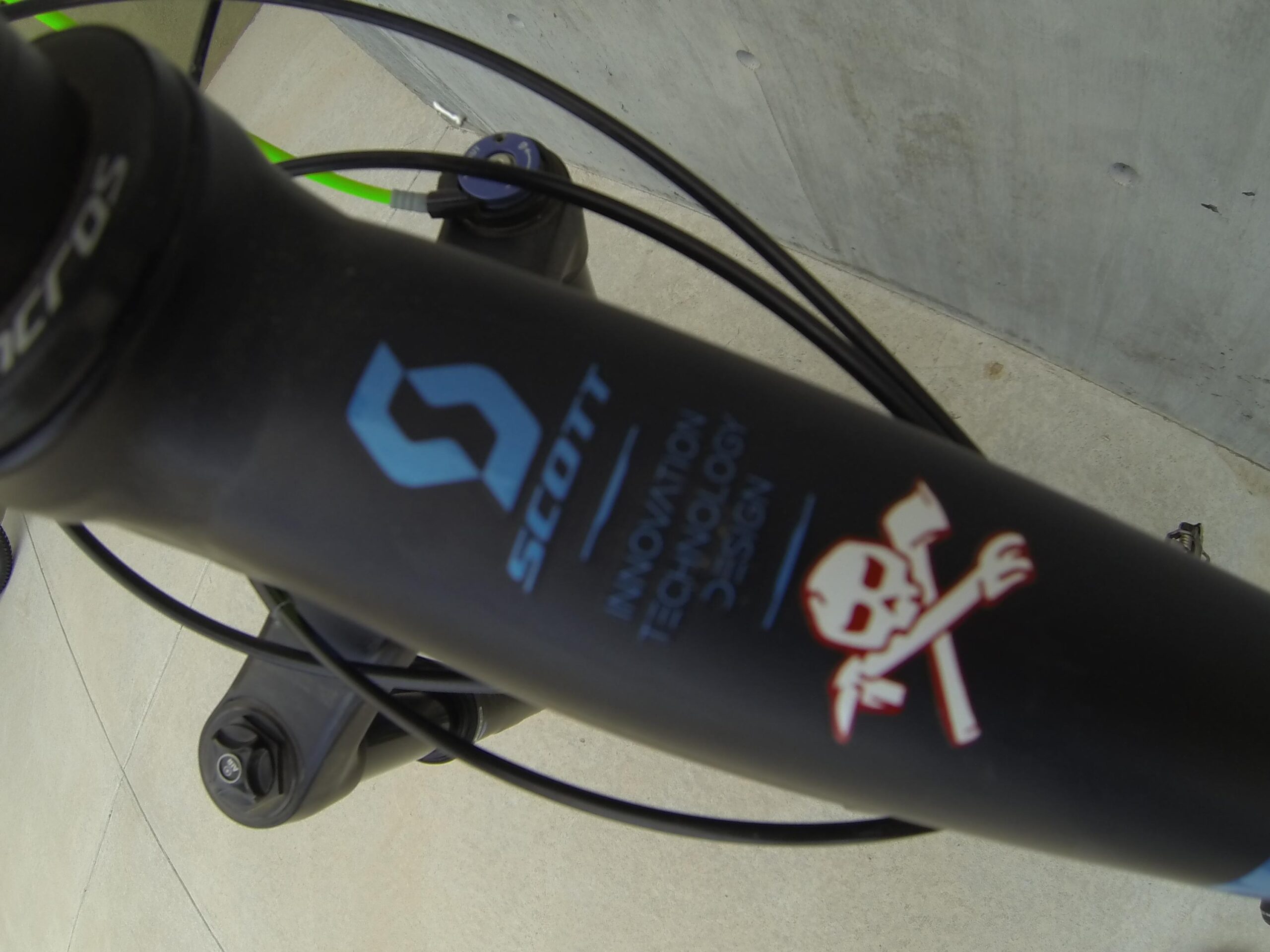 Scott Big Ed: Close-up image of a bike's handlebars showing a logo on the stem that reads "SCOTT" along with a skull and crossbones graphic, against a backdrop of concrete. The handlebars also display wiring and components, emphasizing the bike's design and branding.