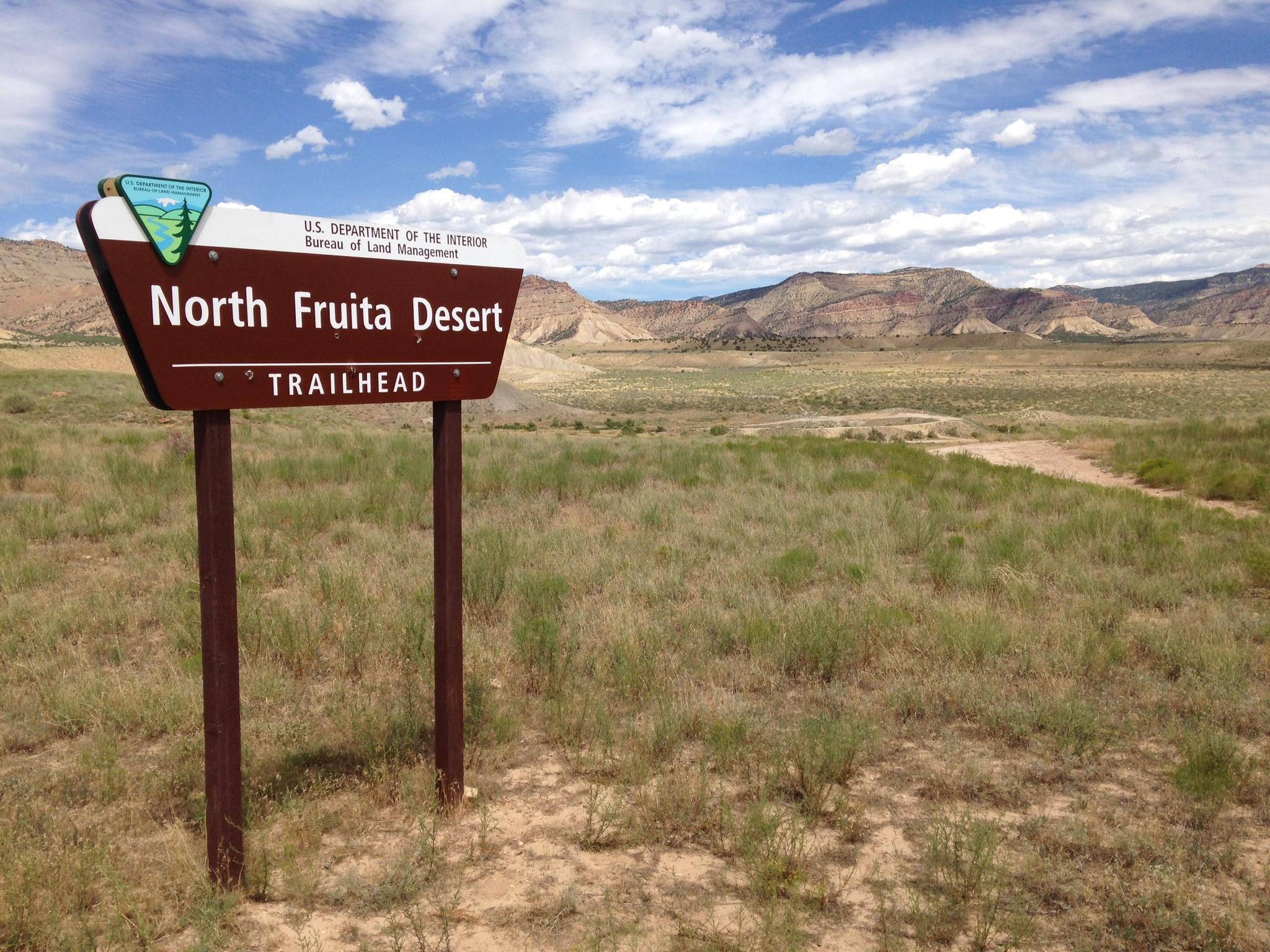 A brown wooden signpost at the North Fruita Desert Trailhead, with mountains and blue sky in the background. The sign features a logo of the U.S. Department of the Interior and indicates the trailhead location. The surrounding landscape includes grassy areas and rocky formations typical of a desert environment. 18 Road Trails / North Fruita Desert mountain bike trail.