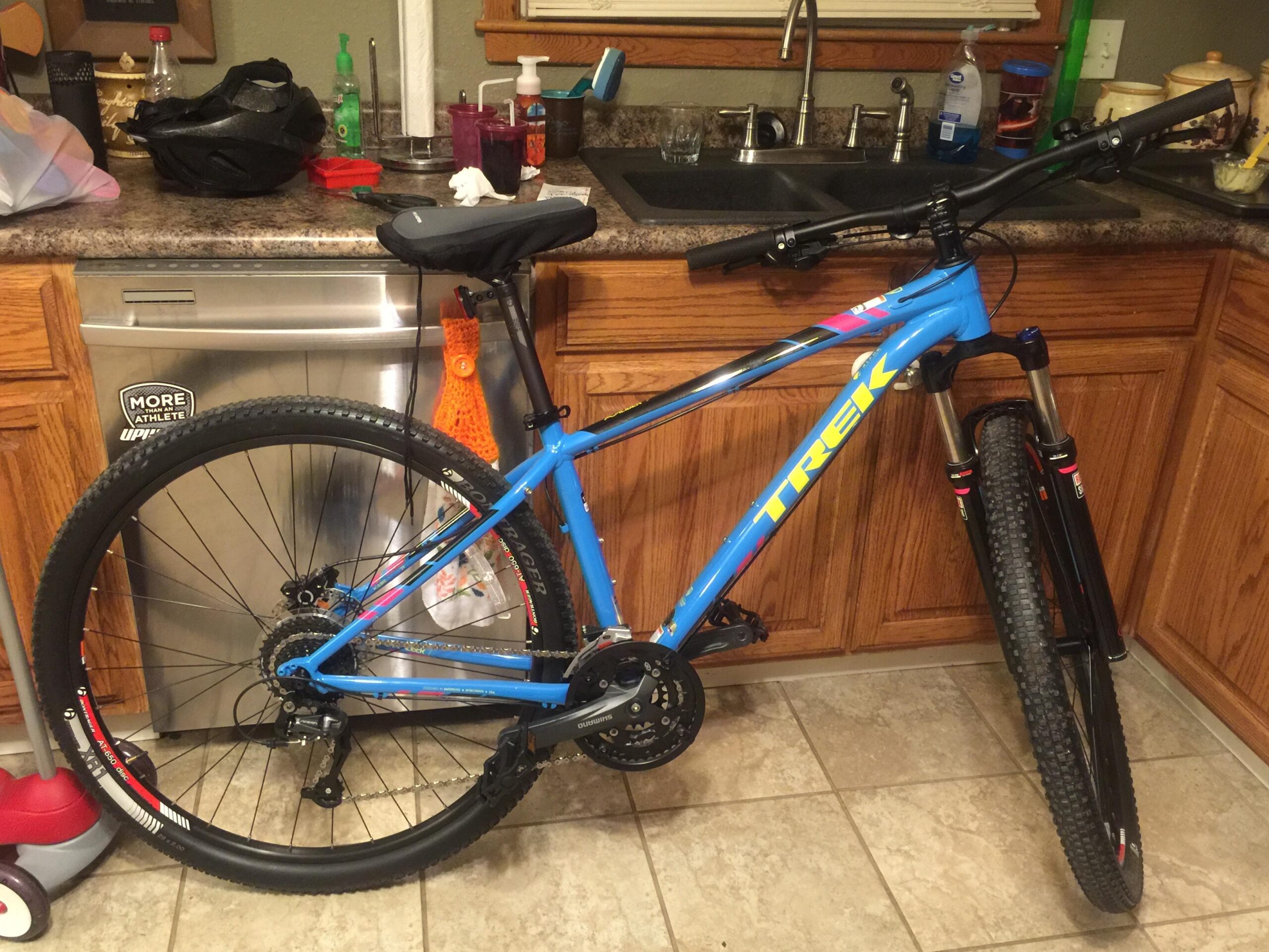 Trek X-Caliber 7: A blue Trek mountain bike leaning against a kitchen countertop, with a helmet and various items on the countertop. The bike features thick tires and a visible drivetrain. In the background, kitchen items include cleaning supplies and dishes on a sink.