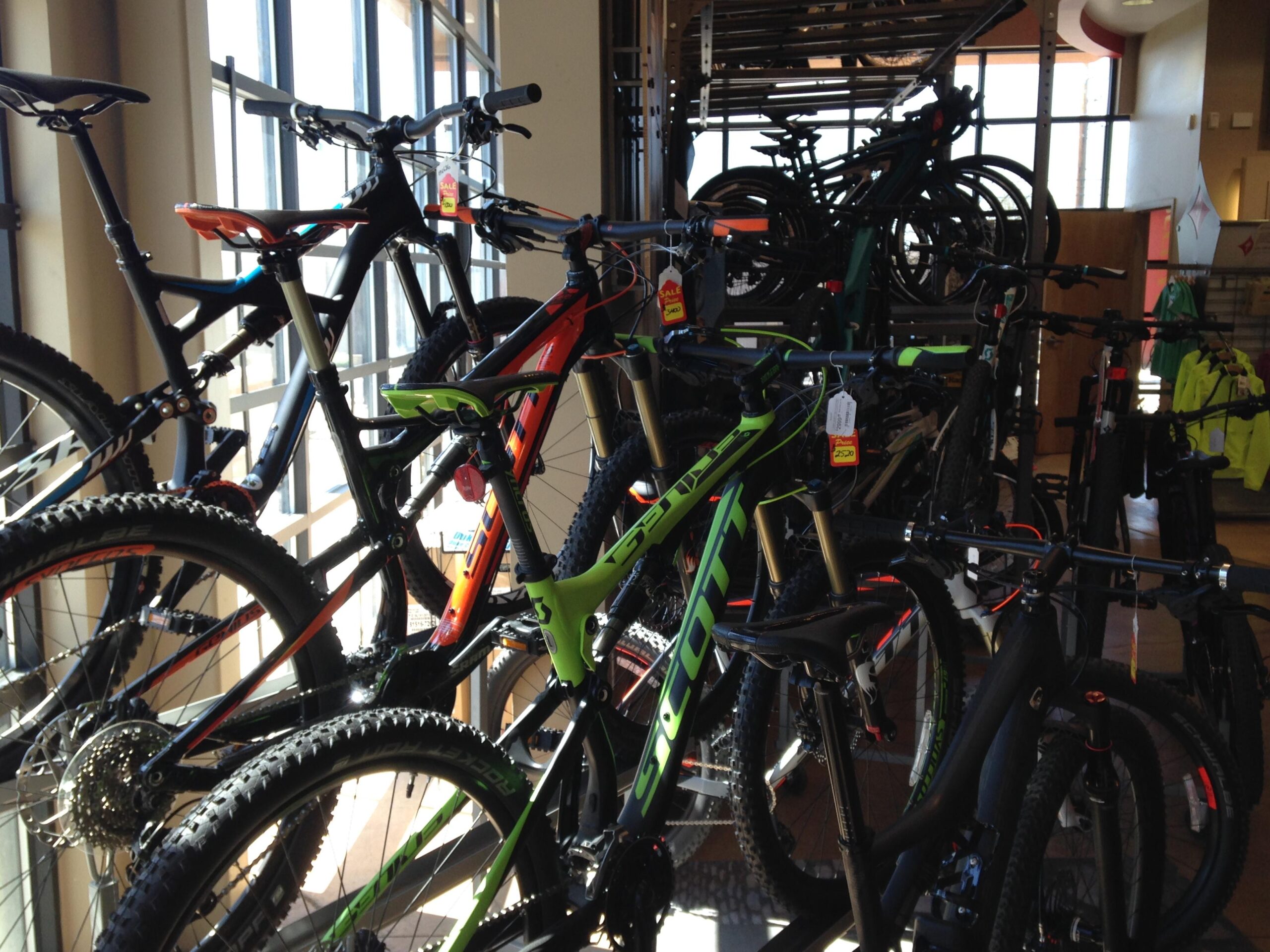 A display of various mountain bikes arranged in a retail store, featuring colorful frames and marked sale tags. The bikes are positioned in front of large windows, allowing natural light to illuminate the scene.