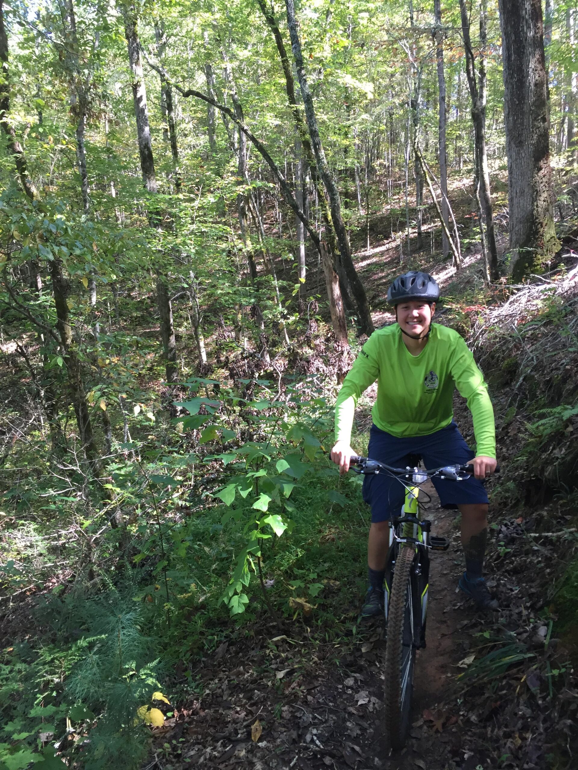 A person wearing a bright green long-sleeve shirt and a black helmet is riding a mountain bike along a narrow dirt trail in a lush forest. Surrounding them are tall trees and green foliage, indicating a scenic outdoor environment. Sunlight filters through the leaves, creating a tranquil atmosphere. Tsali Recreation Area mountain bike trail.