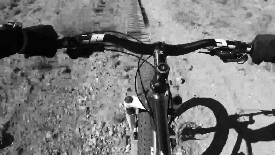 Aerial view of a mountain bike's handlebars, with hands gripping the grips, navigating a dirt trail. The background features a rocky terrain surface, captured in black and white. Goat FatBike Trail mountain bike trail.