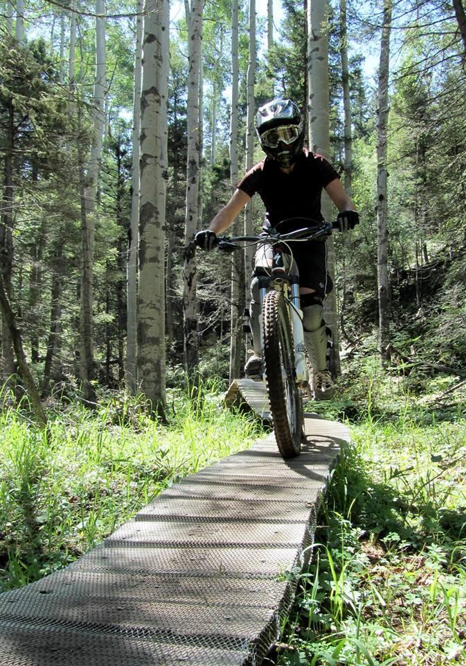 A mountain biker rides across a narrow wooden bridge in a forested area, surrounded by tall trees and greenery. The rider is wearing a helmet and protective gear, showcasing an adventurous biking scenario in nature. Angel Fire Bike Park mountain bike trail.