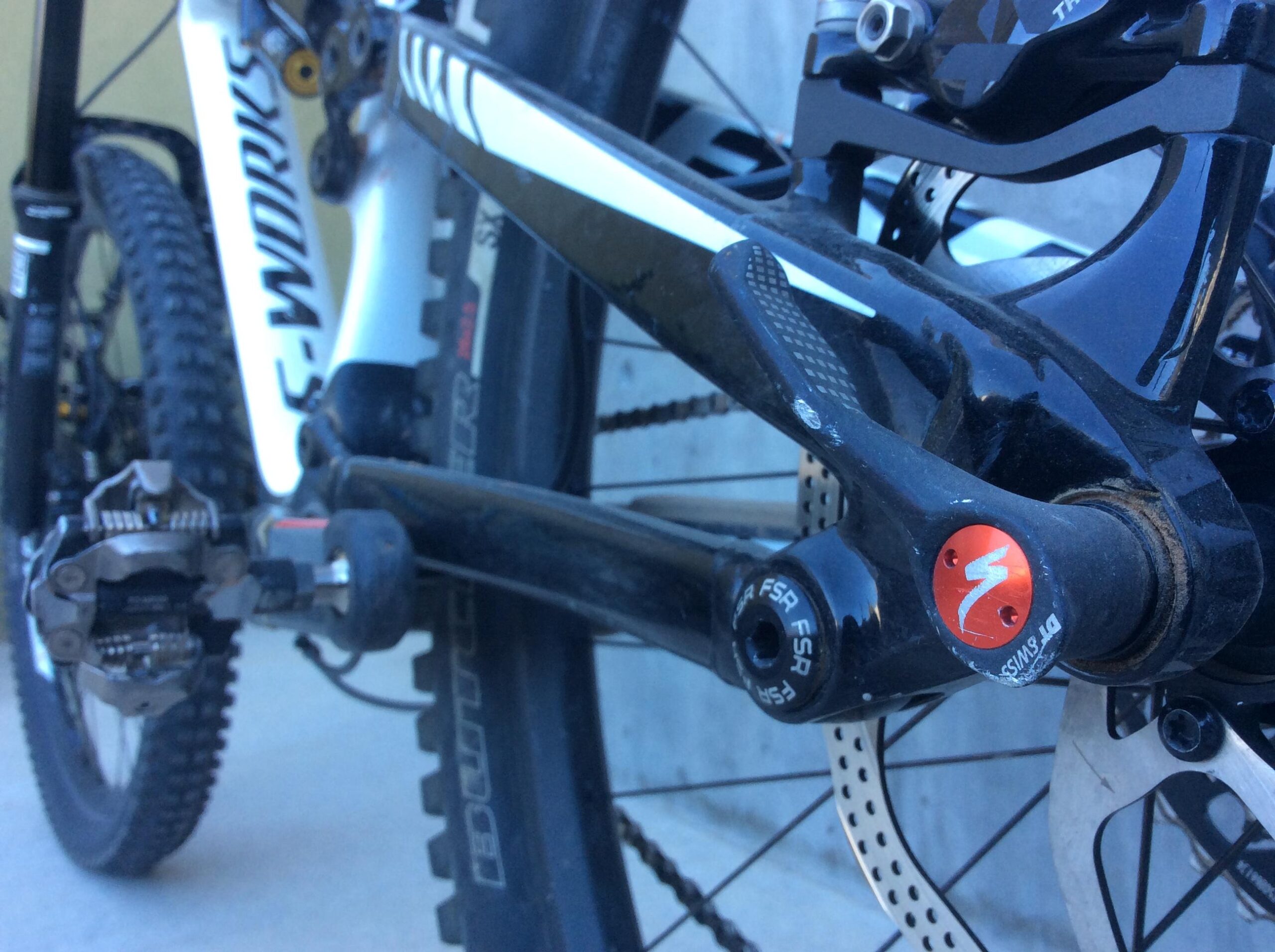 Close-up image of the drivetrain and rear suspension of a mountain bike, showcasing the crank, pedals, and frame details, with a focus on the Specialized branding and components. The background includes a blurred view of the rear wheel and brake disc.