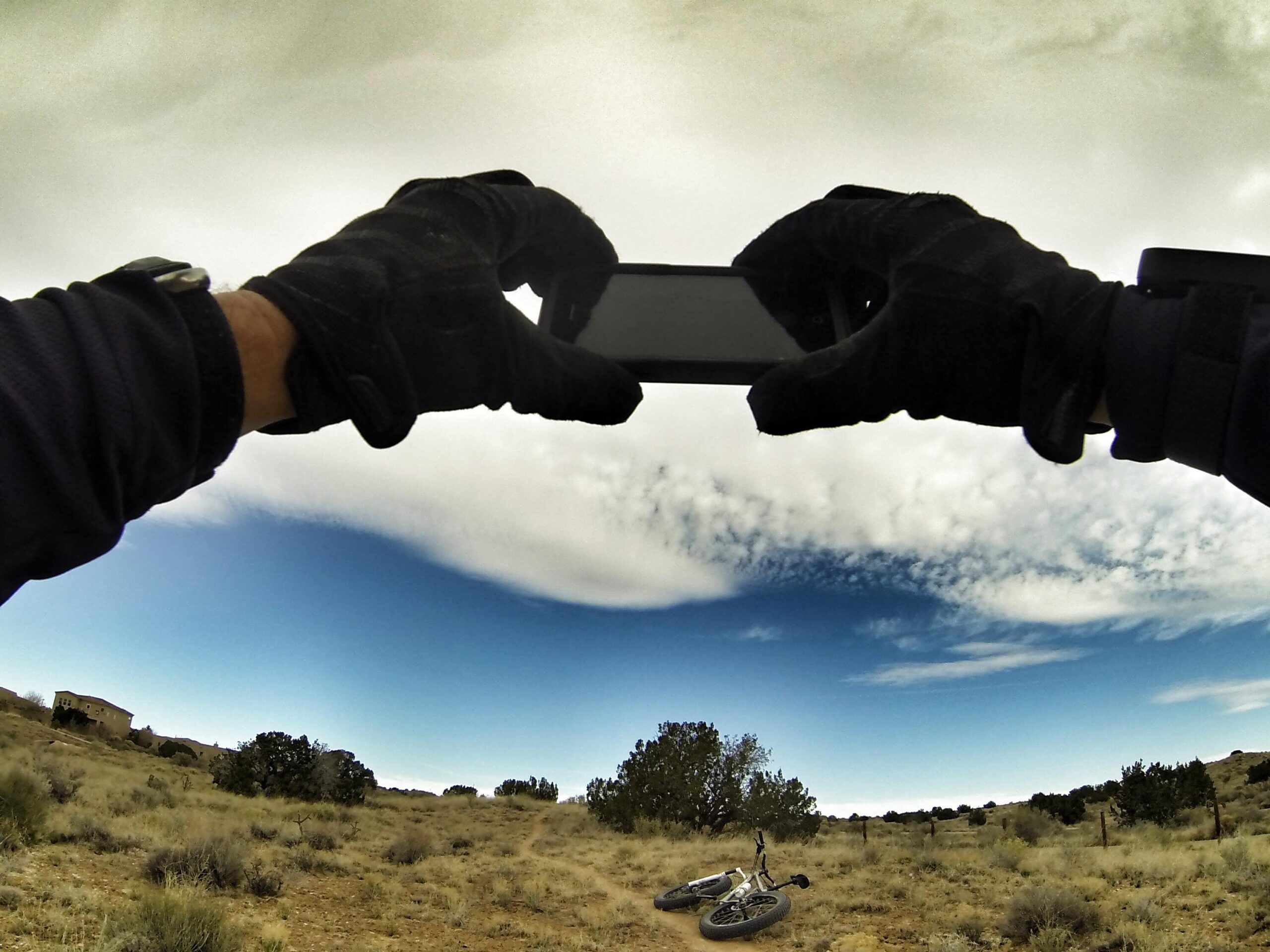 Motobecane Motobecane Bullet Titanium Fat bike: A person's hands wearing black gloves hold a smartphone, capturing a photo of a scenic landscape with a blue sky and scattered clouds. In the foreground, a bicycle rests on the ground amid sparse vegetation and hills in the background.