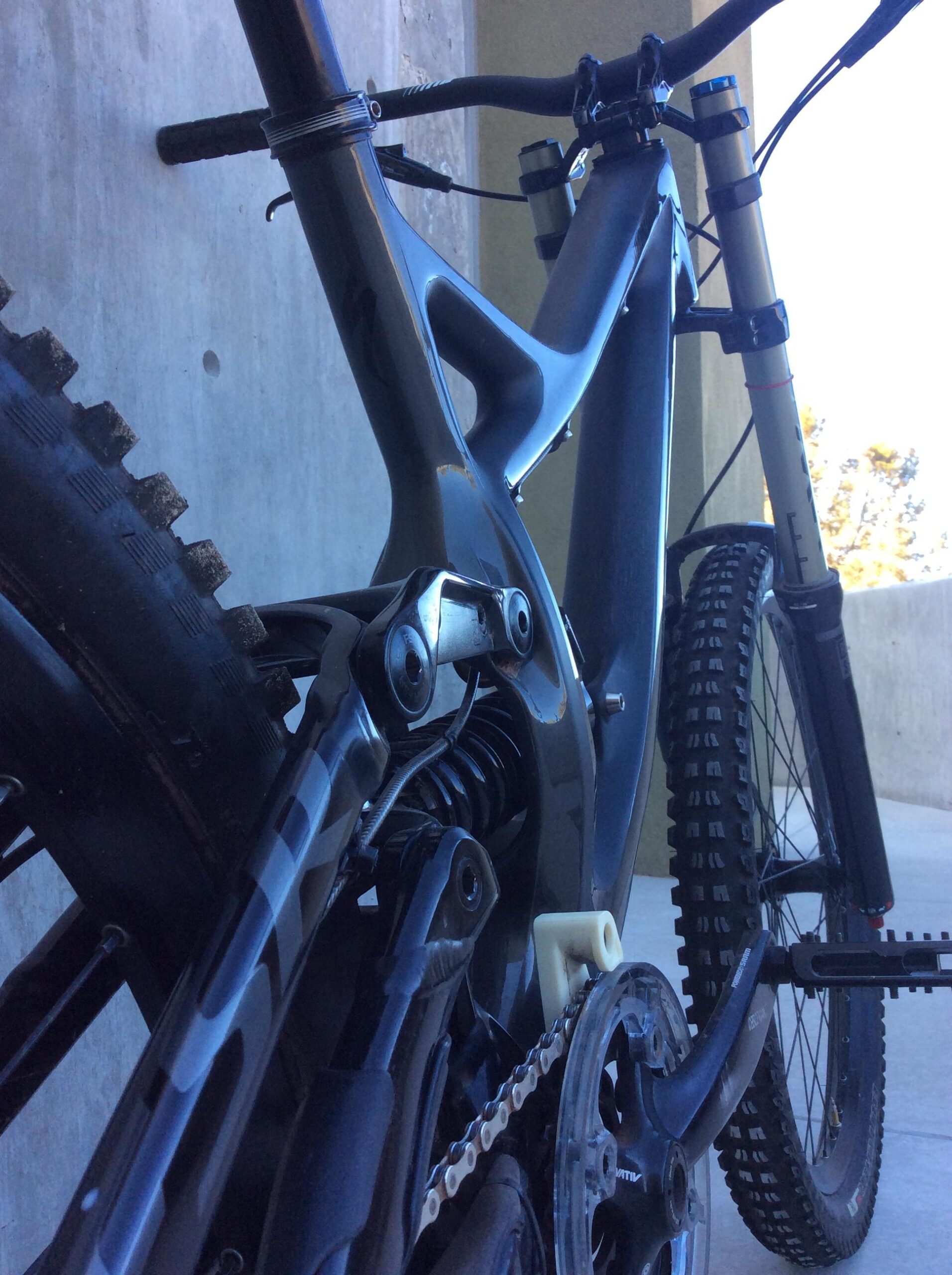 Specialized S-Works Demo 8 Carbon: A close-up view of a mountain bike frame, highlighting the rear suspension system and drivetrain components. The bike is positioned against a concrete wall, with a blurred background showing part of the bike's front fork and wheel. The frame features a sleek, dark design with visible technical details.