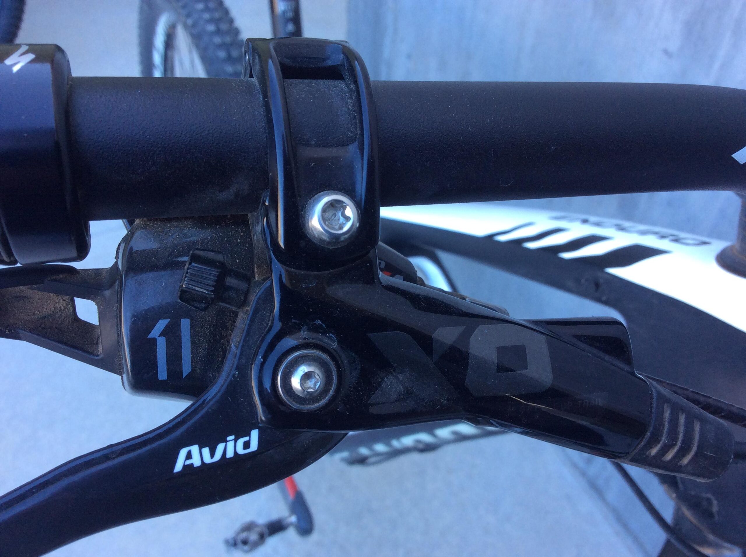 Close-up image of a bicycle's gear shifter, featuring the Avid brand name and the number 11, indicating an 11-speed system. The shifter is black with visible components, including screws and a textured grip, mounted onto the bicycle handlebar. The background is slightly blurred, showcasing part of the bike frame and wheel.