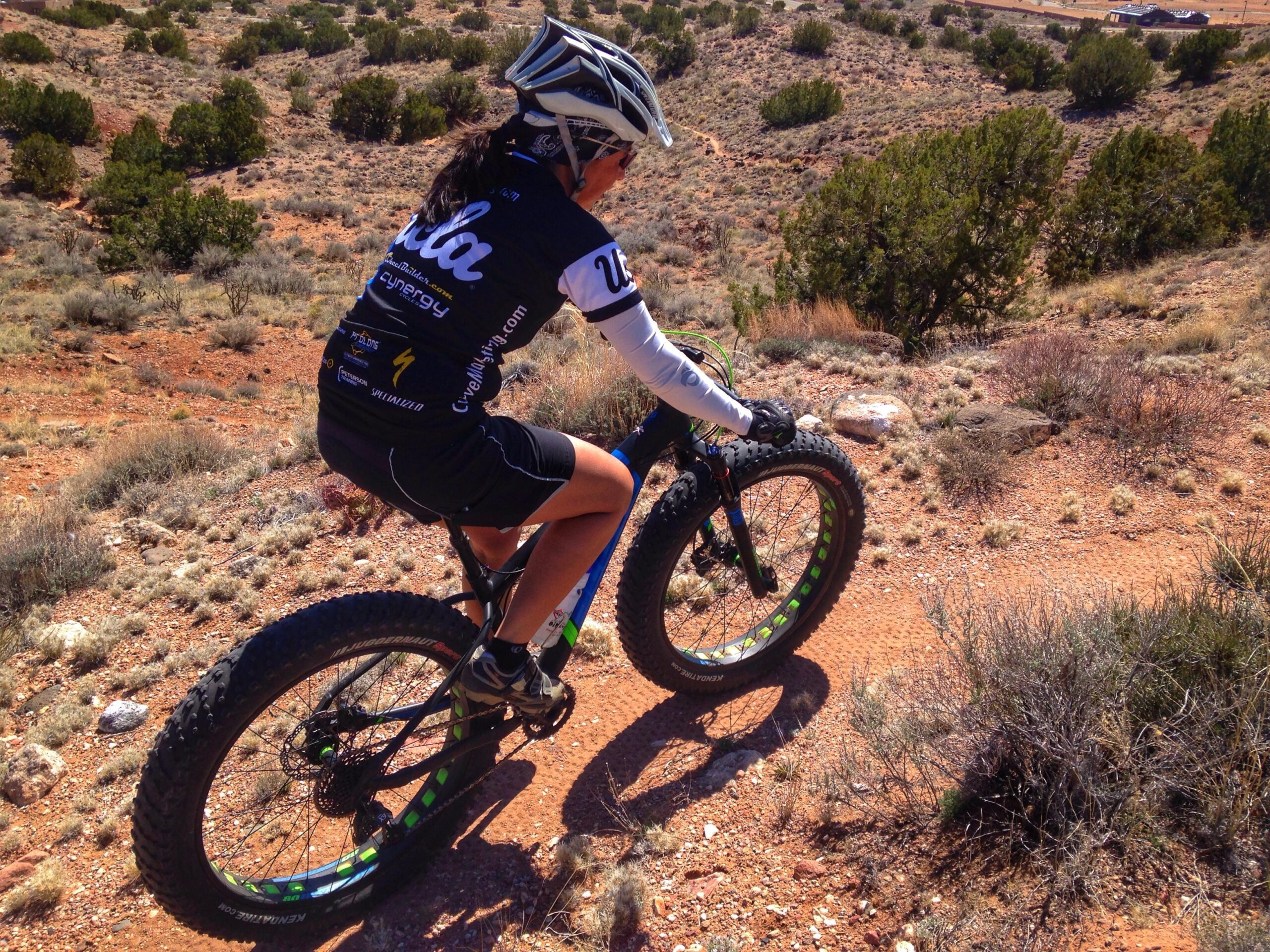 A person riding a fat tire bike on a dirt trail surrounded by shrubs and small trees in a mountainous landscape. The cyclist is wearing a helmet and cycling apparel, and the bike features large, textured tires suitable for rough terrain. Parkway Fatbike trail mountain bike trail.