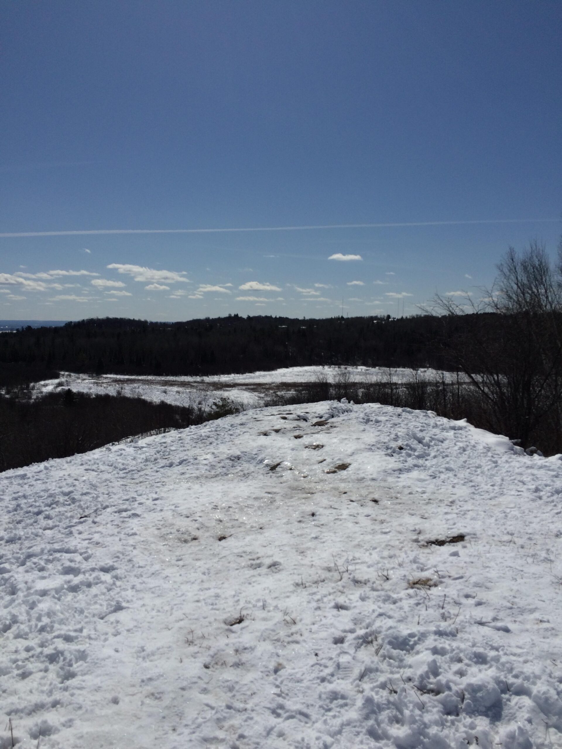 A snow-covered hilltop with a clear blue sky and scattered clouds, overlooking a winter landscape of rolling hills and trees in the distance. Hartley Park mountain bike trail.