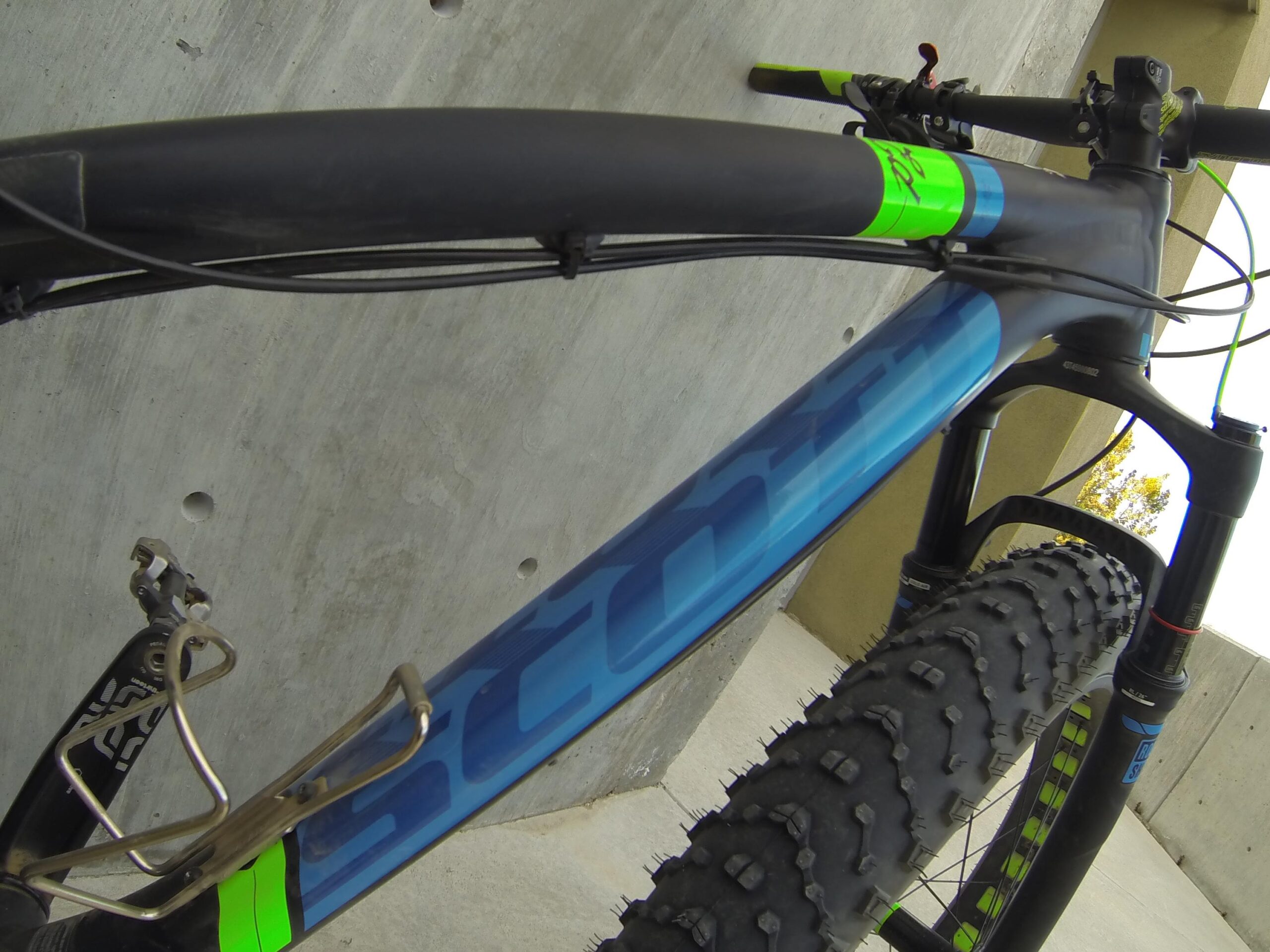 Scott Big Ed: Close-up view of a mountain bike frame featuring a blue design with bright green accents. The image shows the handlebars and the front tire, which has a rugged tread suitable for off-road terrain. The background consists of a concrete surface and a wall, indicating an outdoor setting.