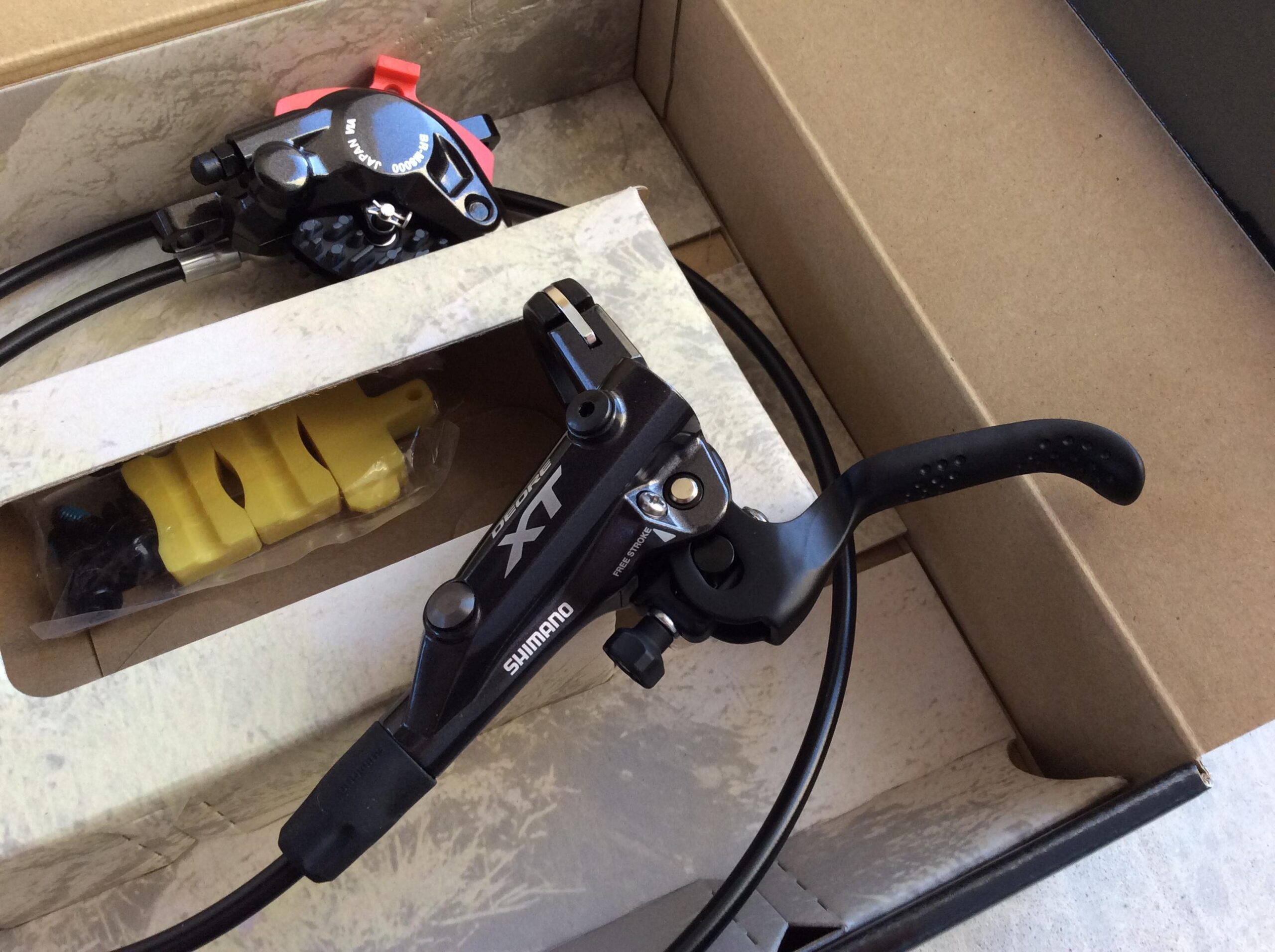 Alt text: "A Shimano XT hydraulic brake lever and caliper set, displayed in a cardboard box. The box contains yellow brake pads and screws, alongside the black brake components and their connecting cables."