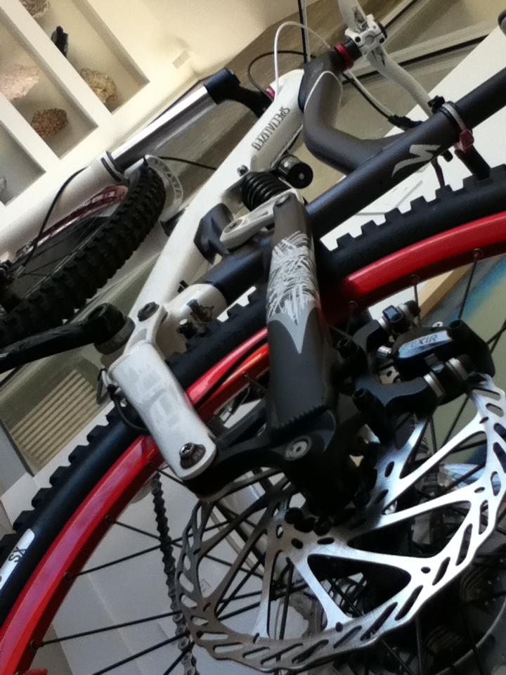 A close-up view of two mountain bikes stacked on top of each other, showcasing the handlebars, tires, and brake disc components. The image highlights the contrasting colors of the bikes, with one featuring white and gray elements and the other with red accents. The background is softly blurred, emphasizing the bikes.