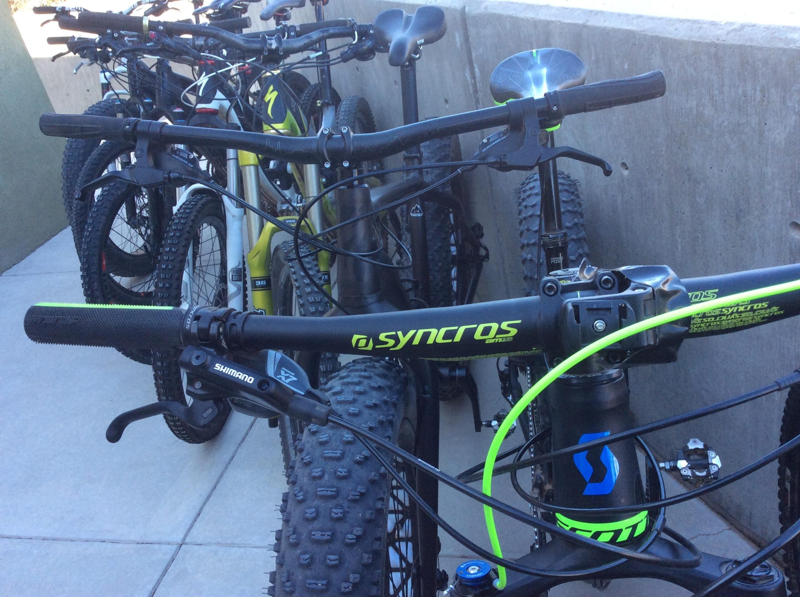 A close-up view of several mountain bikes parked closely together. The image prominently displays the handlebars and components of a bike with a green and black design, featuring a "Syncros" logo. Other bikes can be seen in the background, showcasing a variety of styles and colors. The setting appears to be a concrete area, suggesting a bike rack or storage space.