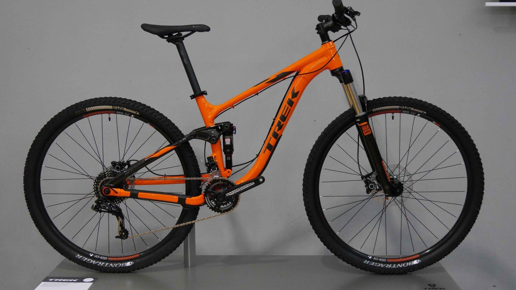 Trek Fuel EX 5 29: A bright orange mountain bike with a durable frame, featuring wide tires and front suspension, displayed against a neutral gray background. The bike is positioned on a stand, highlighting its design and components.