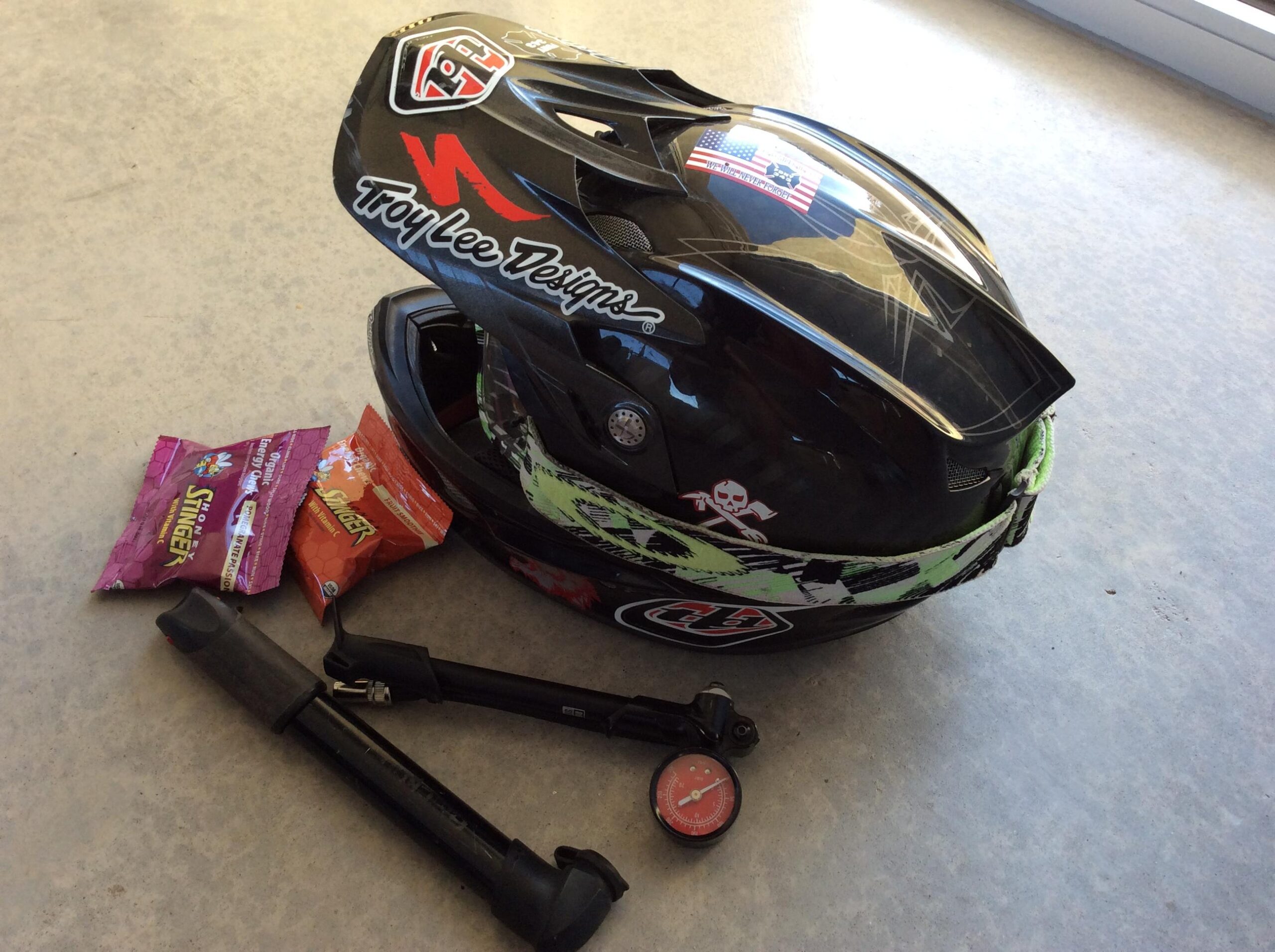 A black motorcycle helmet with colorful designs, including logos from Troy Lee Designs and a small American flag, is placed on a concrete surface next to two energy bar packages and a bike pump with a pressure gauge.