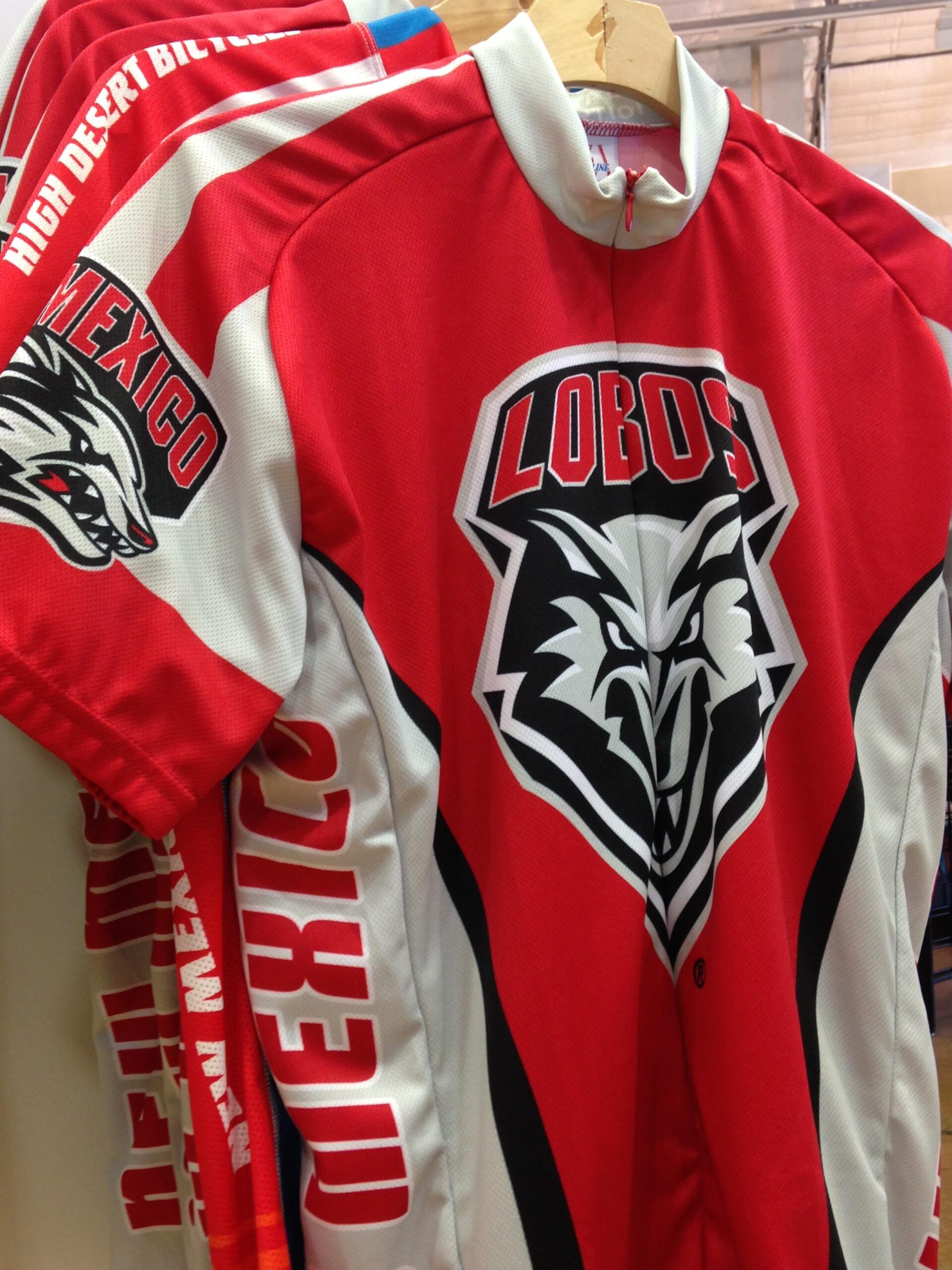 A close-up view of a red and white cycling jersey featuring the logo of the Lobos, with the word "Mexico" printed in bold letters on the sleeve. The jersey is displayed on a hanger, showcasing its sporty design and branding.