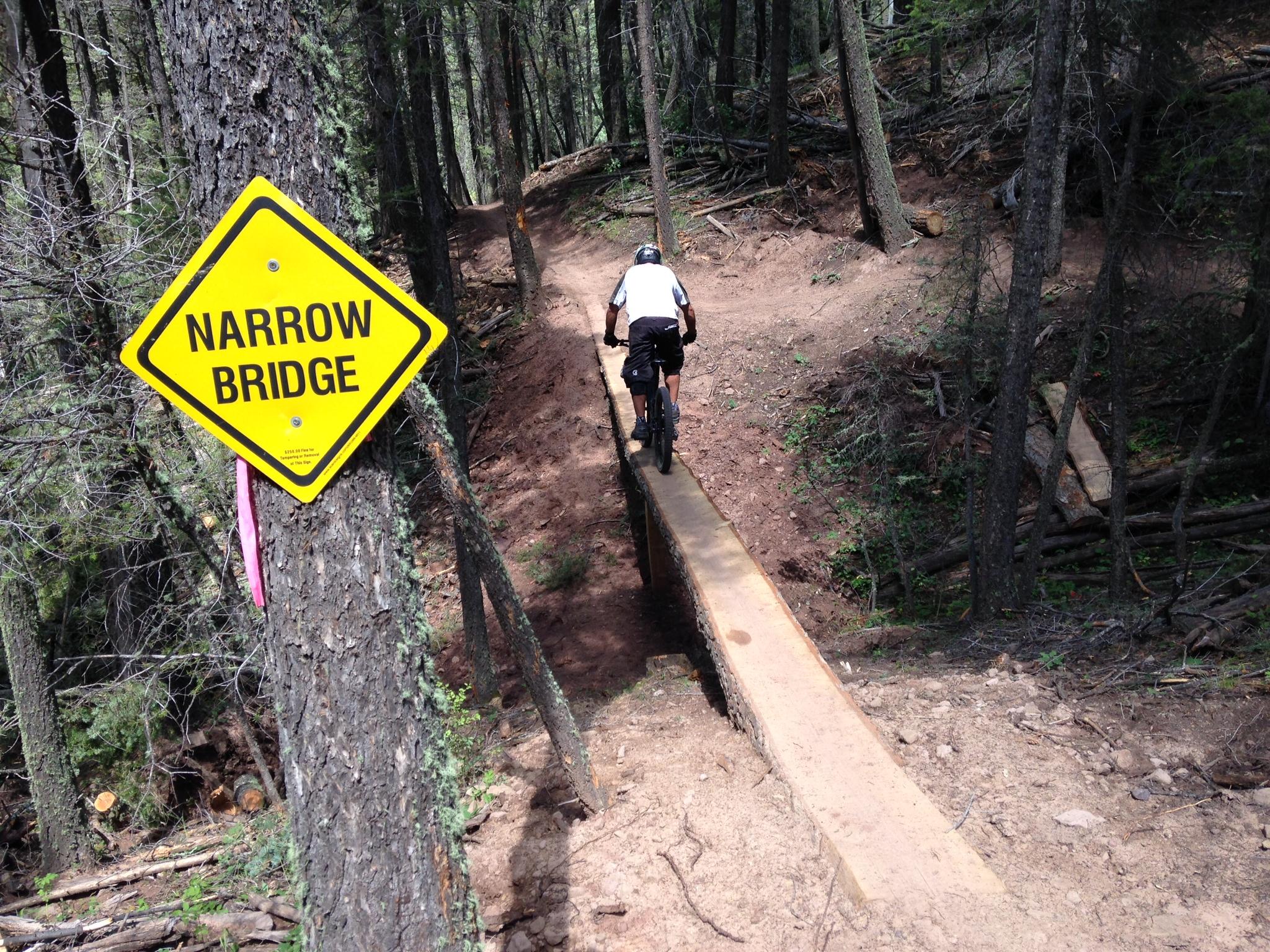 Specialized Enduro S-Works 650b: A cyclist riding a mountain bike across a narrow wooden bridge in a wooded area, with a "Narrow Bridge" warning sign attached to a tree nearby. The trail is lined with trees and uneven terrain.