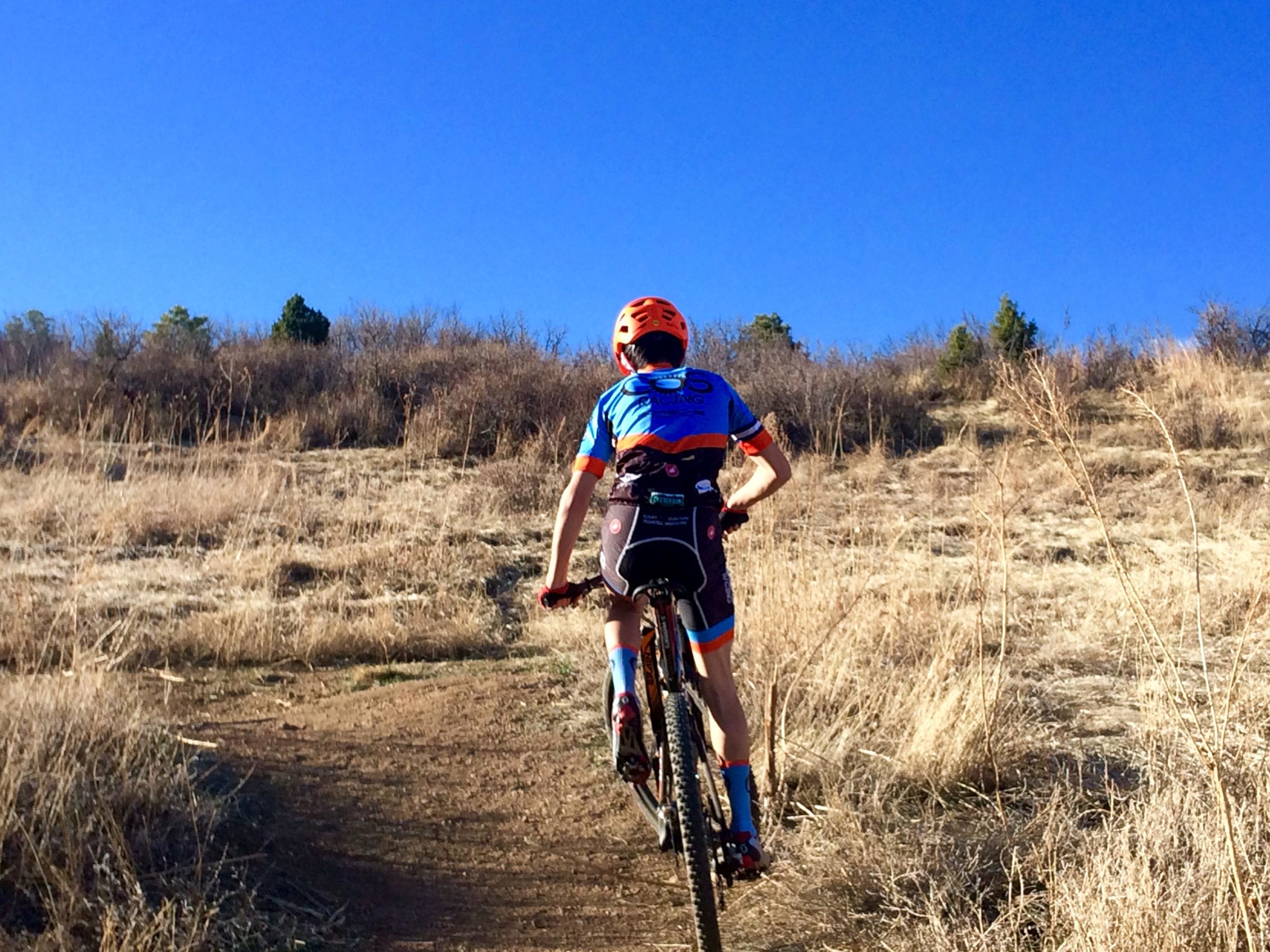 A young cyclist wearing a bright orange helmet and a blue and orange biking jersey rides up a dirt trail leading to a grassy hillside, under a clear blue sky. Stratton Open Space / The Chutes mountain bike trail.