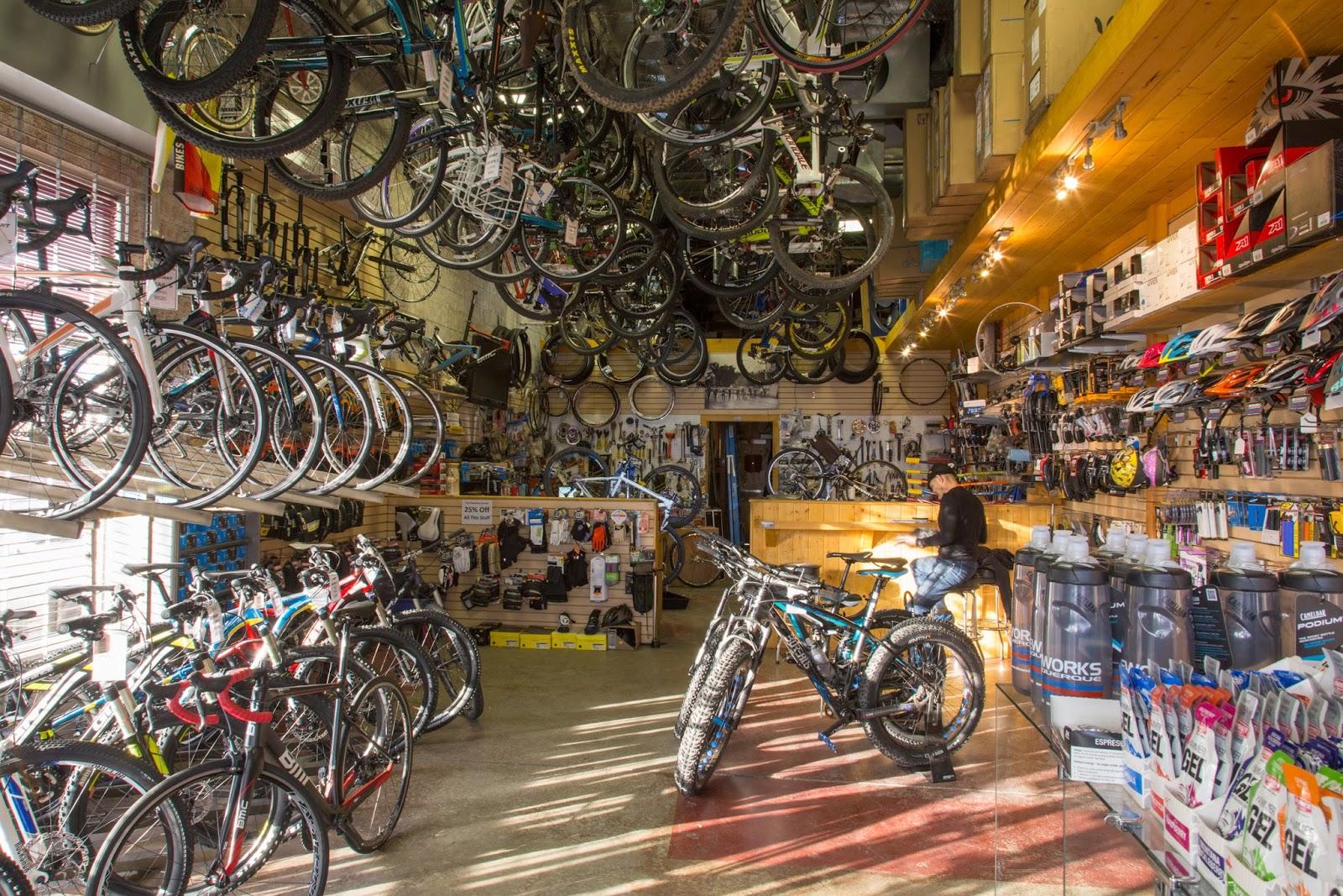 A well-stocked bicycle shop featuring a variety of bicycles hanging from the ceiling and displayed on the floor. The interior includes shelves filled with bike accessories, tools, and merchandise, and a customer sitting at a service counter in the background. The lighting is bright, highlighting the array of products available.