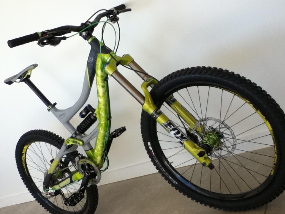 A mountain bike with a distinctive green and gray design, featuring a suspension system and oversized tires, is leaning against a light-colored wall. The bike has handlebars with grips and includes a prominent "FOX" logo on the front fork.