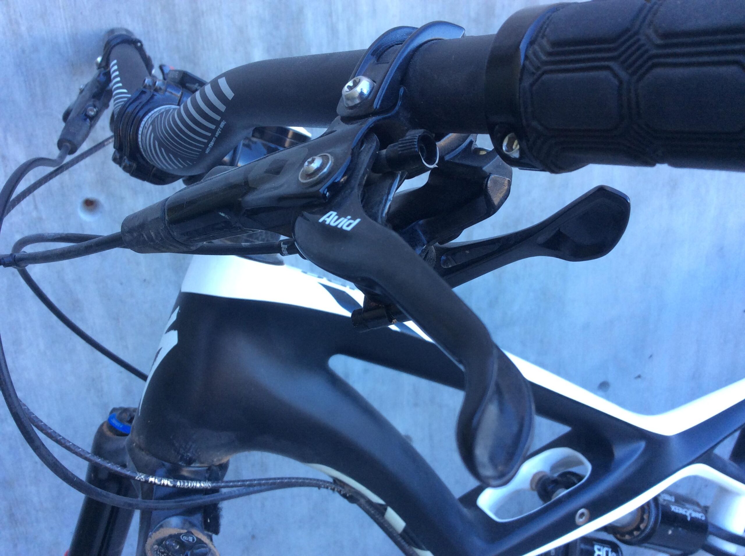 Close-up view of a bicycle's handlebars, featuring black brake levers, grips, and cables against a textured concrete background. The brake levers are labeled "Avid," indicating their brand, and the handlebars have a sleek, modern design typical of mountain or performance bikes.