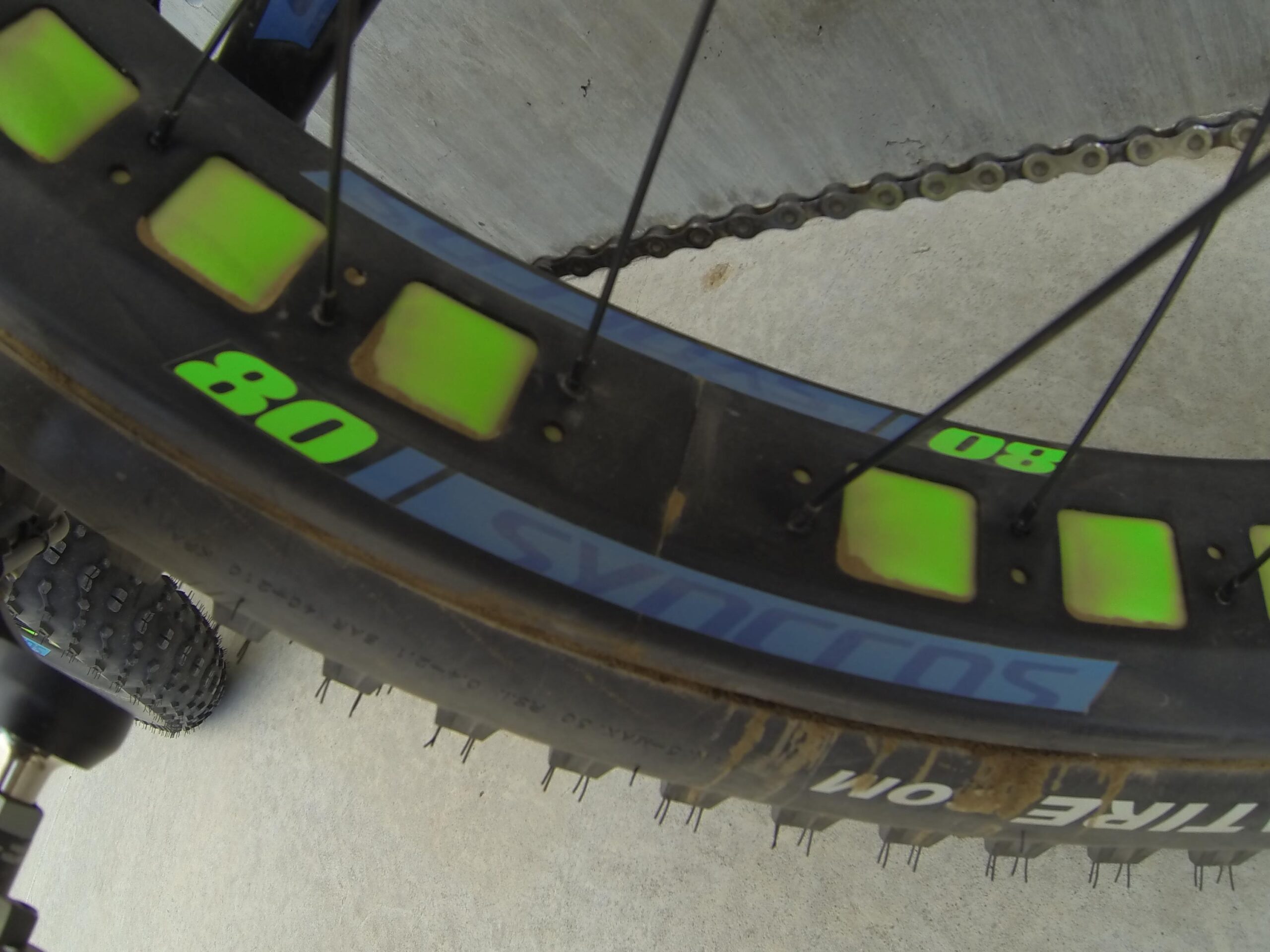 Scott Big Ed: A close-up view of a bicycle wheel with a focus on the rim, featuring neon green accents and the brand name "Syncros." The wheel shows signs of use, with some dirt and wear visible, and the tire has a knobby texture. The background is a concrete surface.