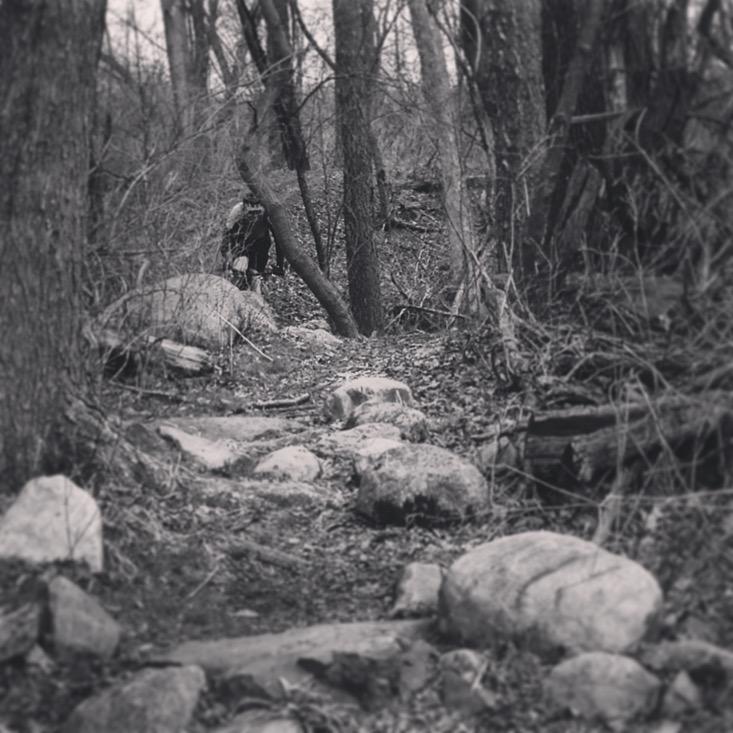 A black and white photo of a wooded path featuring large stones along the trail. Sparse trees with bare branches surround the path, creating a moody and natural setting. In the distance, a figure is partially visible among the trees, emphasizing the sense of exploration in the wilderness. The Dump mountain bike trail.