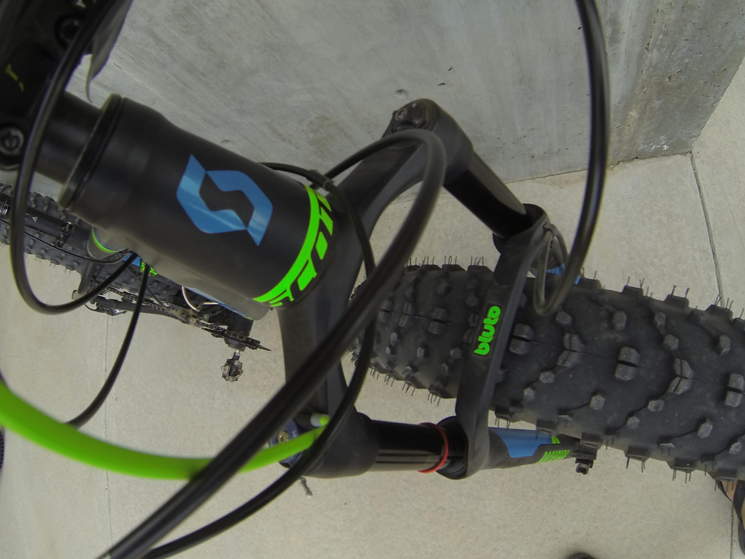 Scott Big Ed: An overhead view of a mountain bike's front fork and tire, highlighting the bike's branding details and the textured surface of the tire. The image features the bike's suspension components, cables, and a portion of the ground surface.