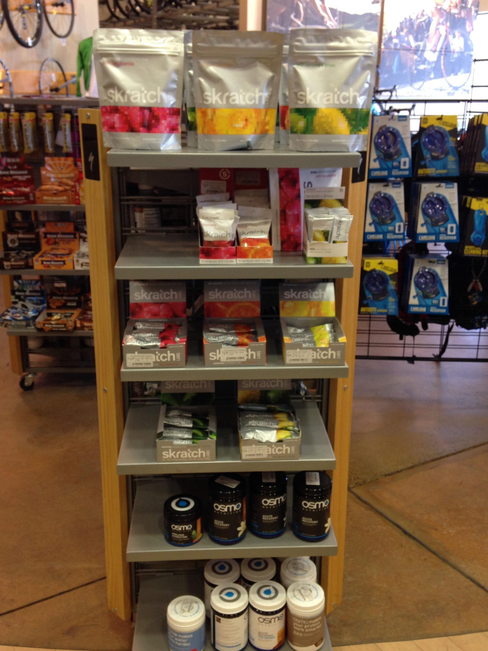 A display shelf in a retail store showcasing various sports nutrition products, including Skratch Labs items in colorful pouches and Osmo containers. The shelf is well-organized with different flavors and types of energy products, set against a backdrop of cycling gear.
