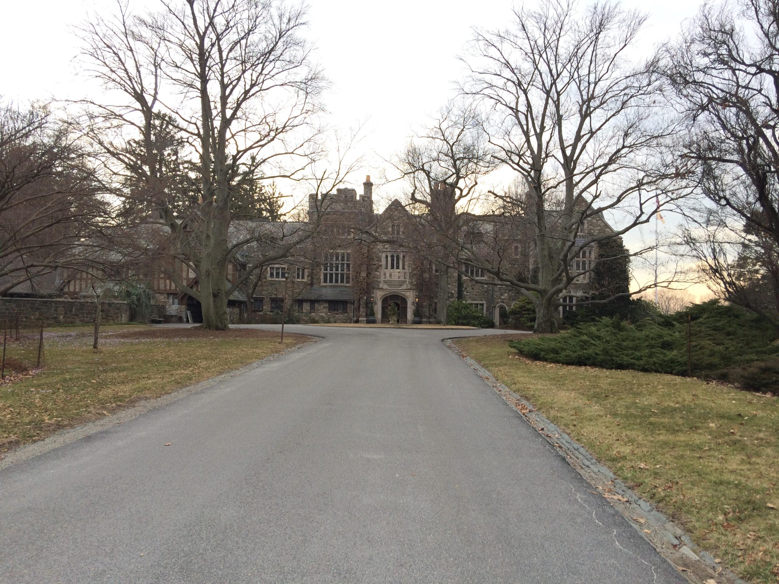 A winding driveway leads to a large, historic stone mansion surrounded by bare trees and shrubs. The sky is overcast, hinting at an early evening. The mansion features a mix of architectural styles with prominent windows and intricate stonework. Ringwood Skylands Manor mountain bike trail.