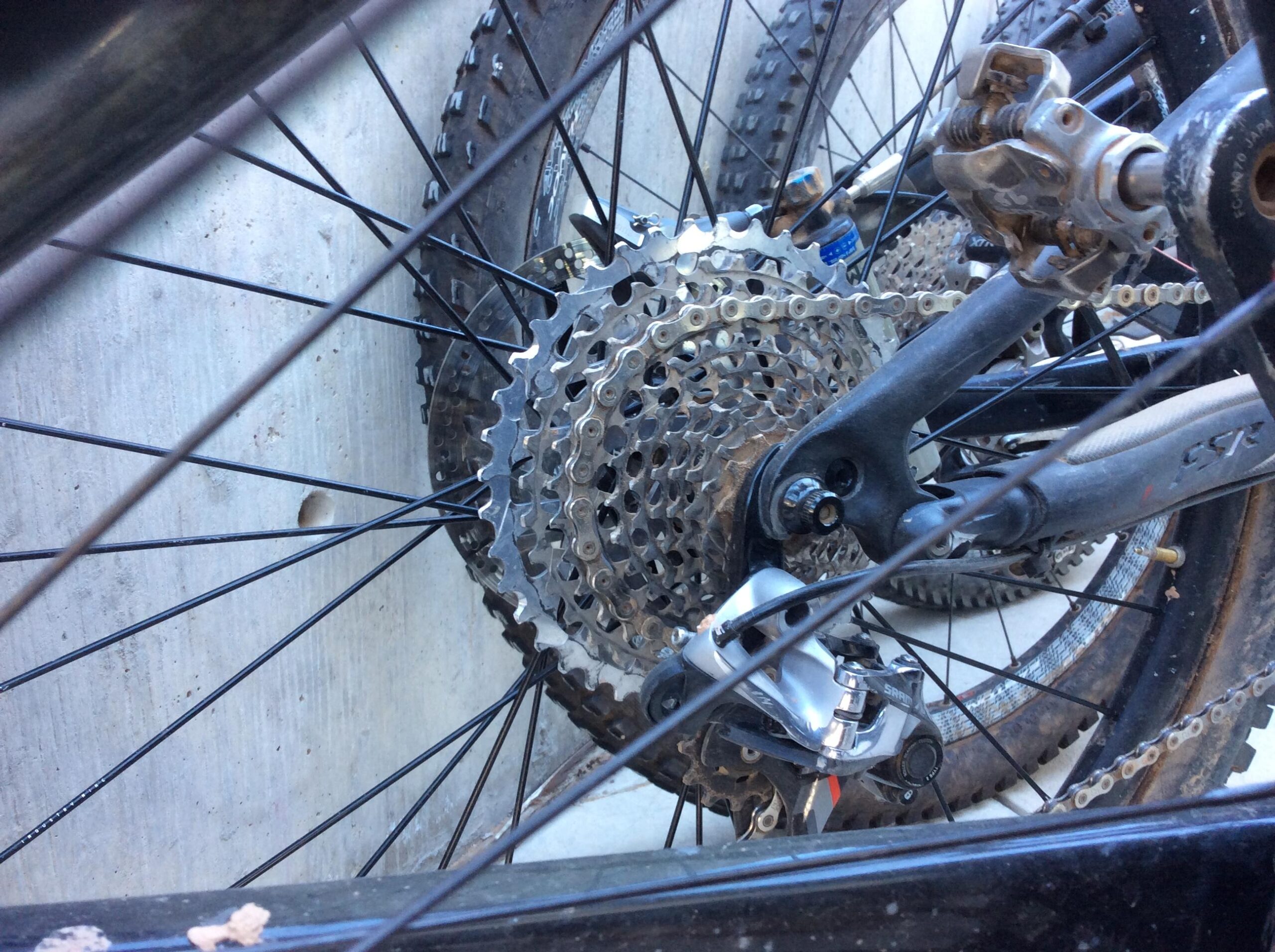 Close-up image of a bicycle rear gear system, showcasing the cassette, chain, and derailleur amidst bicycle spokes and nearby wheels. The background features a textured concrete surface.