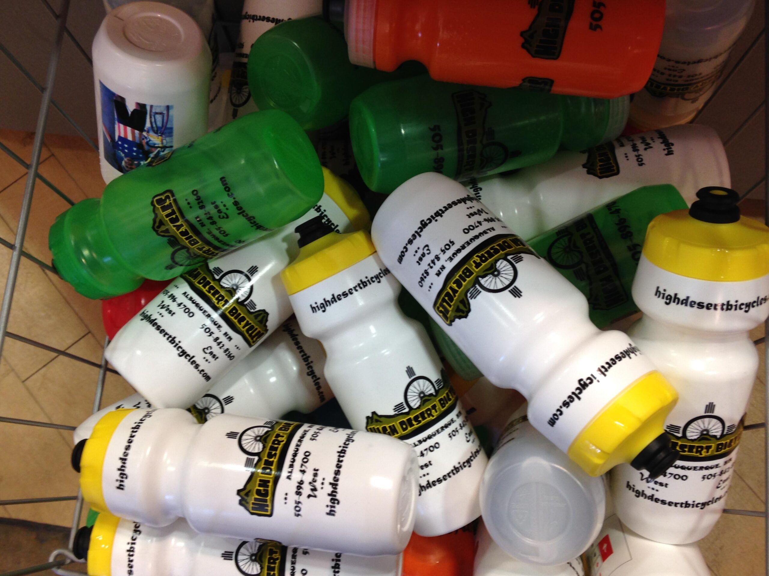 A colorful assortment of bicycle water bottles in a wire basket, featuring various designs including green, red, orange, and white bottles. Some bottles display the logo and contact information for "High Desert Bicycles." The arrangement shows the bottles stacked together in a casual, disorganized fashion.