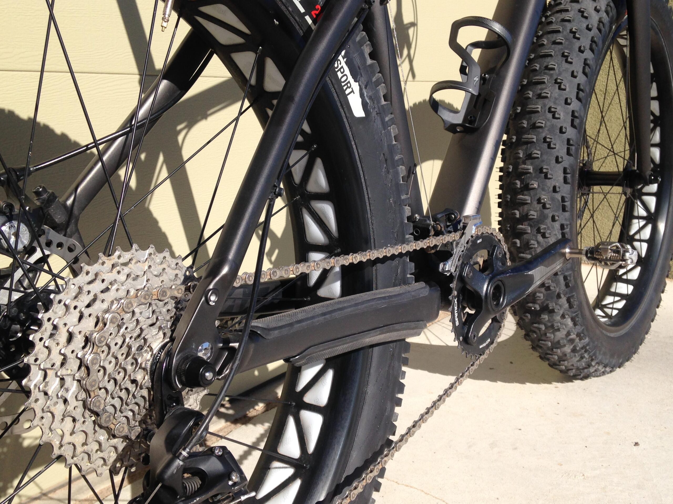 Close-up of the rear gears and chain of a mountain bike, featuring a large cassette and wide tire. The bike is positioned against a light-colored wall, showcasing the mechanical components, including the crankset and chain.