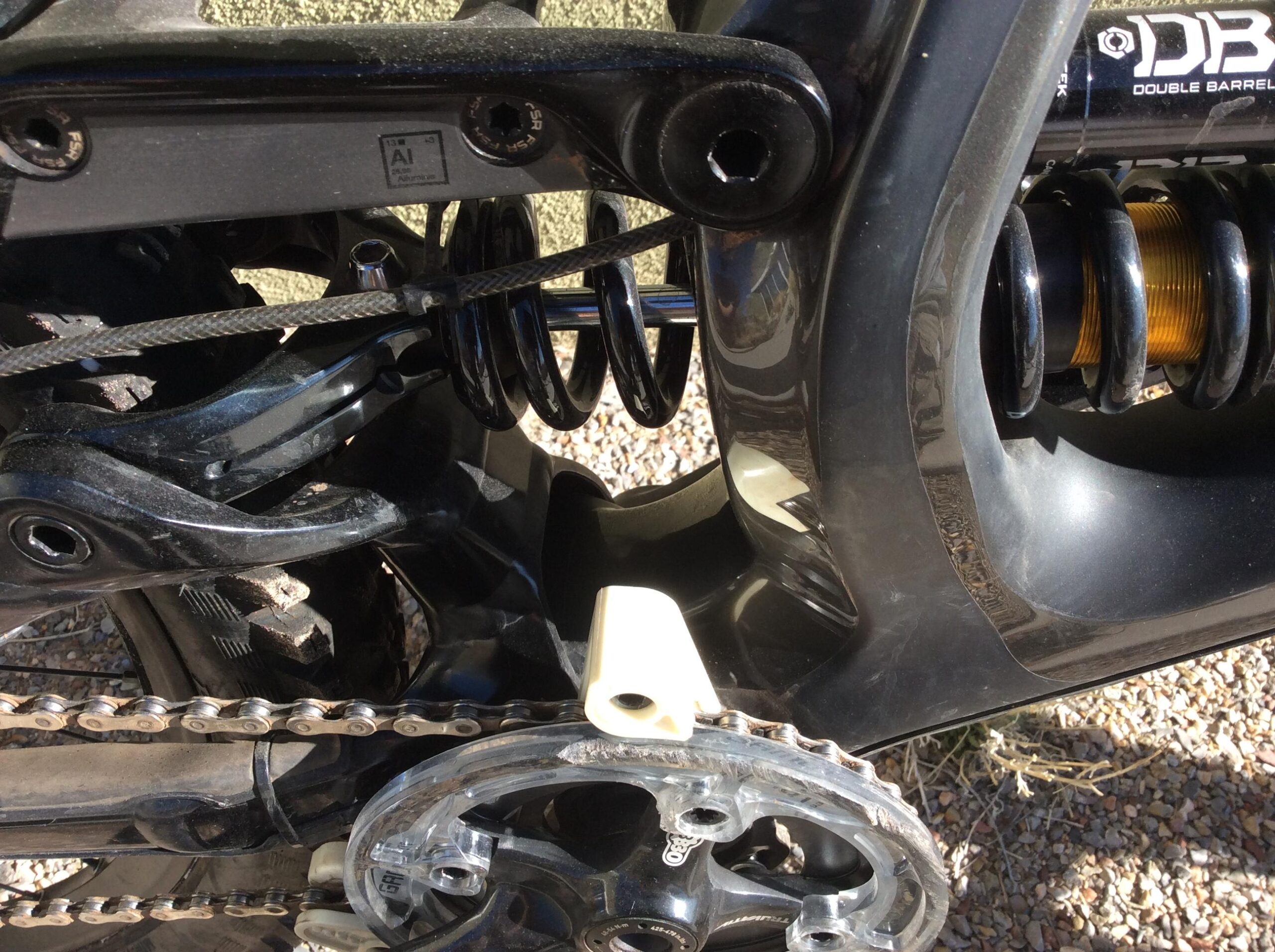 Specialized S-Works Demo 8: Close-up view of a bicycle's rear suspension system, featuring a black shock absorber and coil spring, along with a portion of the chain and crankset. The image highlights mechanical details and construction materials against a textured background.