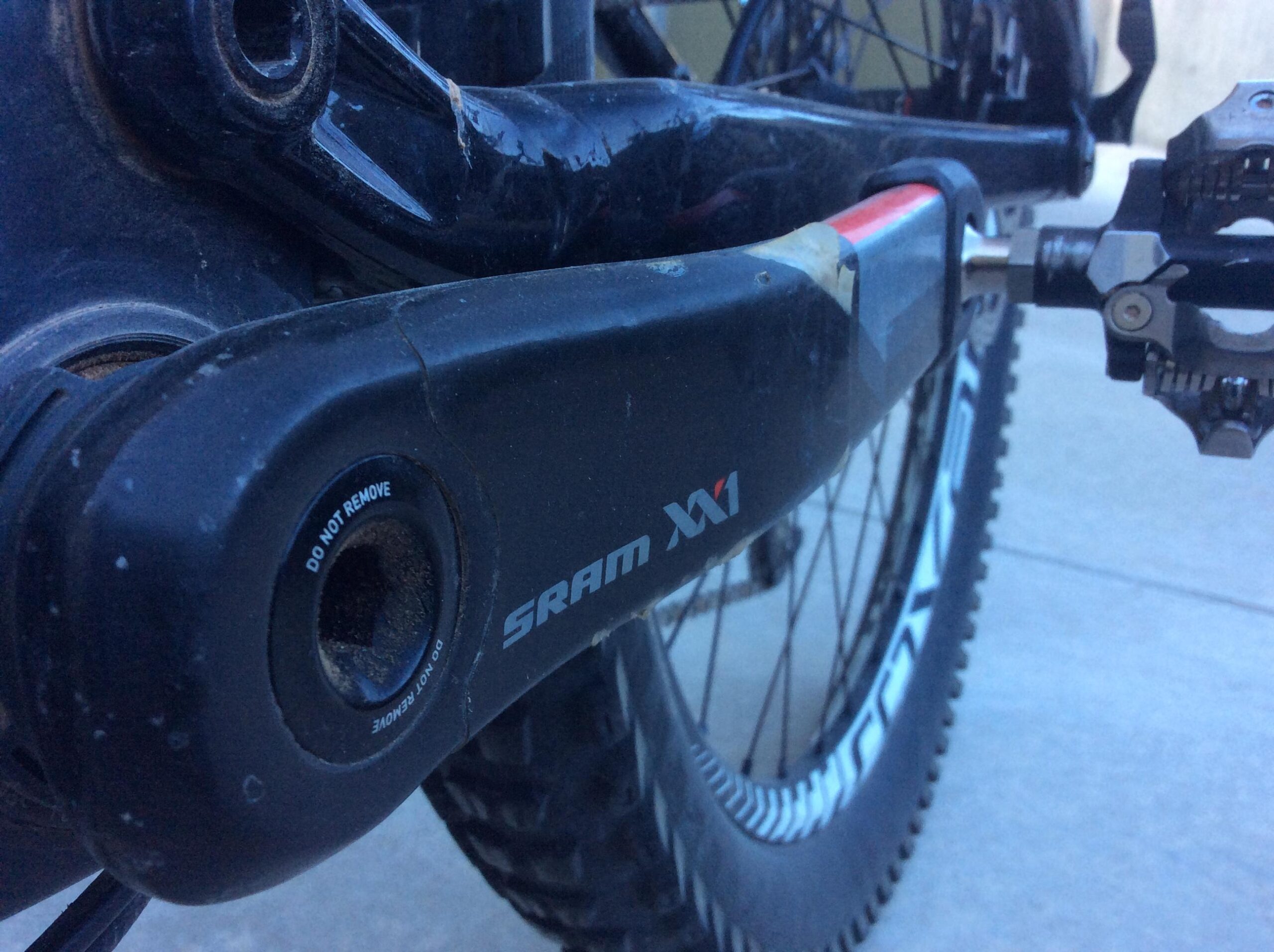 Close-up of a SRAM XX1 bicycle crankset, showing the crank arm, pedal attachment, and visible bearings. The image highlights the logo and the "DO NOT REMOVE" marking on the crank arm, with a blurred background of the bike's rear wheel and ground surface.