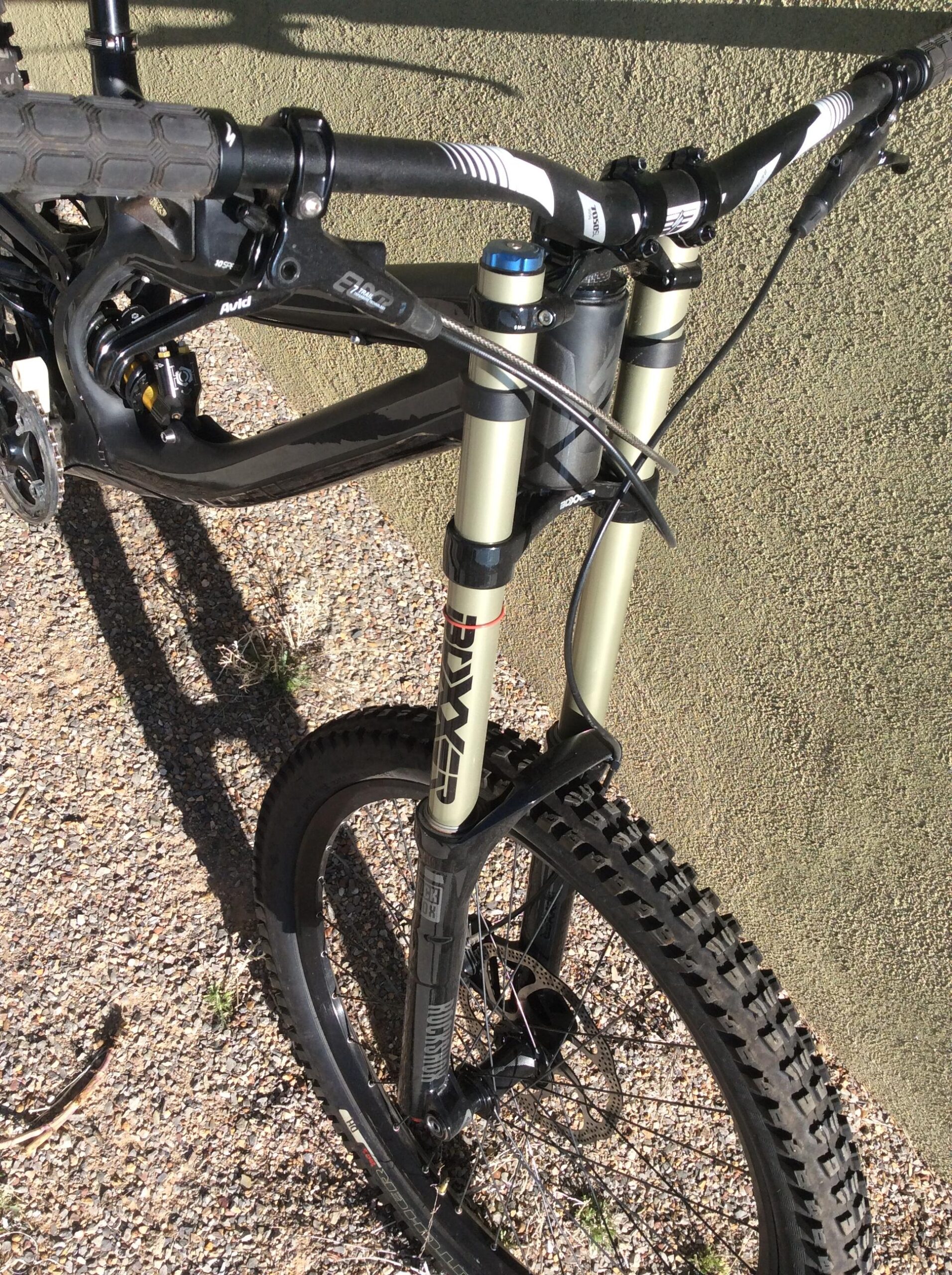 Specialized S-Works Demo 8: Close-up view of a mountain bike's front suspension and wheel, showing the adjustable fork, handlebars, and brake lever. The bike is positioned on gravel, with a textured wall in the background.