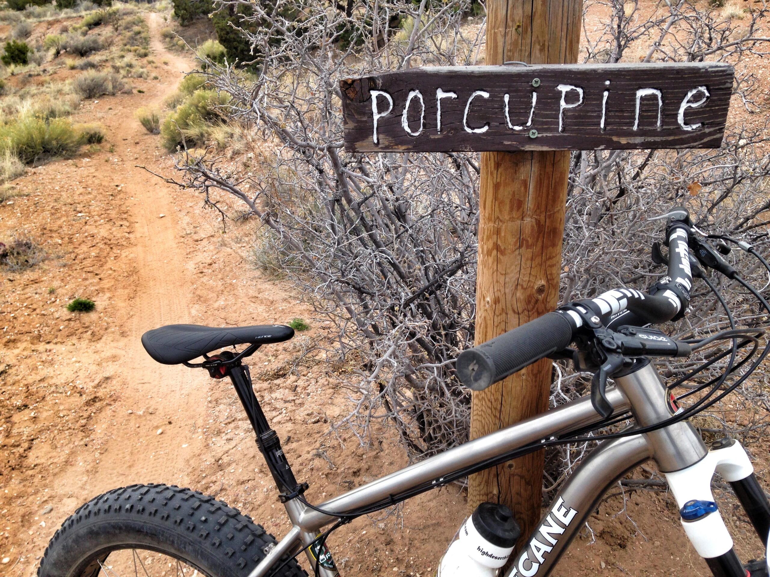 A mountain bike with fat tires parked next to a wooden sign that reads "Porcupine," with a dirt trail winding in the background through a desert landscape featuring sparse vegetation and rocky terrain. Parkway Fatbike trail mountain bike trail.
