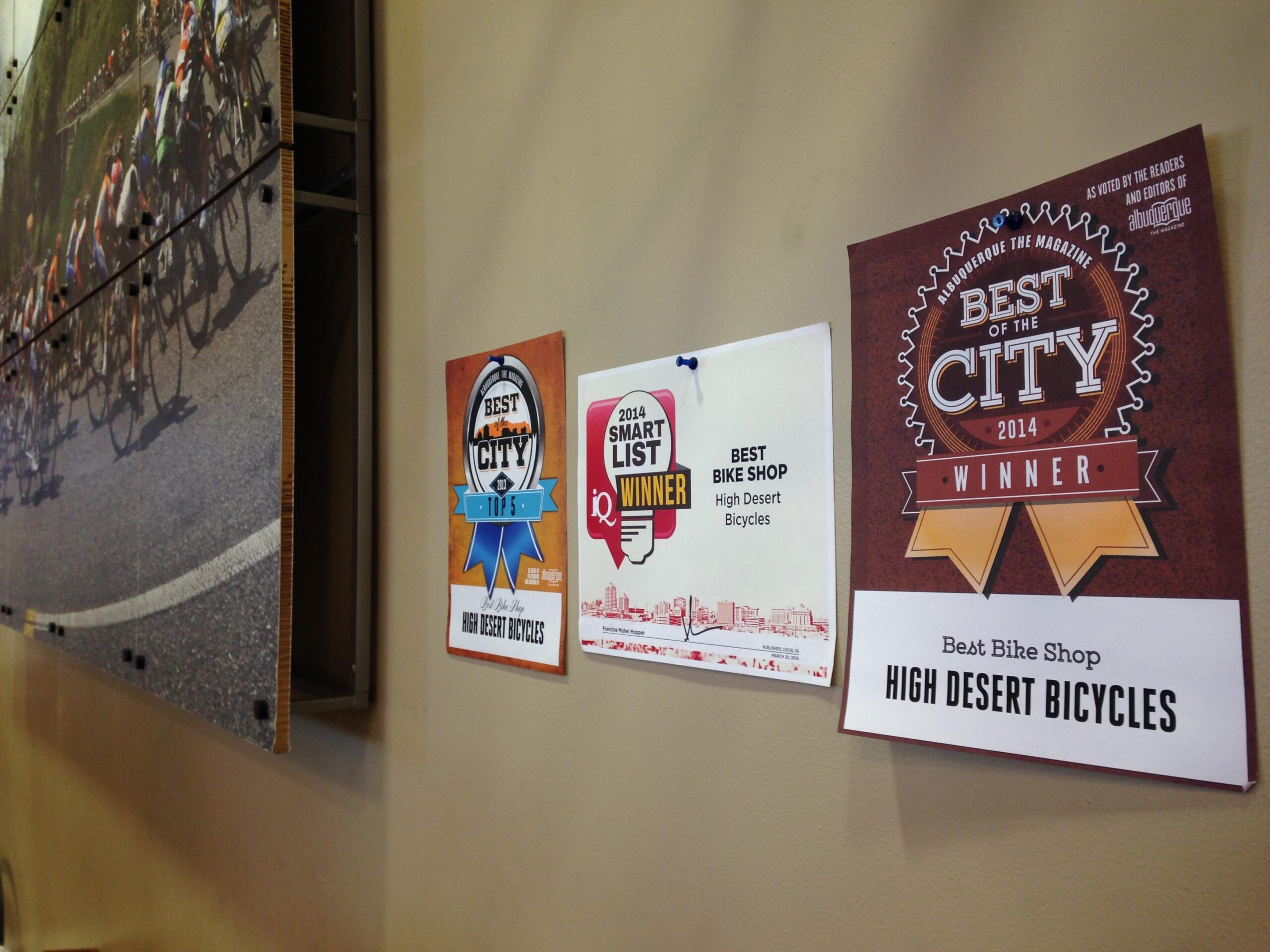 A wall display featuring awards for "Best of the City" from 2014, highlighting High Desert Bicycles as the best bike shop, along with a "Smart List Winner" certificate. In the background, there is a large photo depicting cyclists racing on a road.