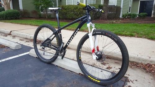 Niner EMD 9: A mountain bike with a black frame, yellow grips, and white front fork rests against a curb in a parking lot. The bike features thick tires suitable for off-road cycling. In the background, there are green trees and residential buildings.