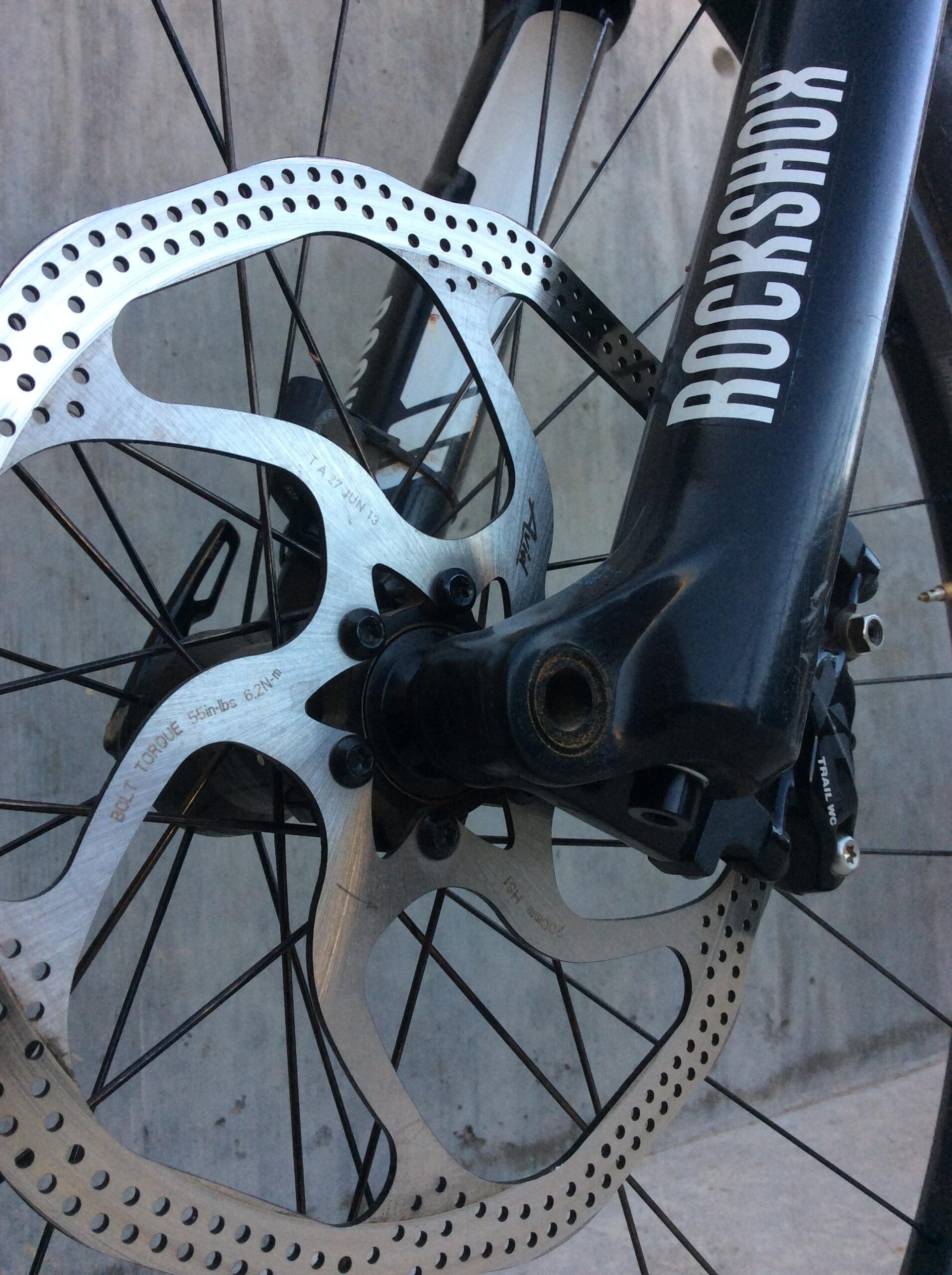 Close-up image of a bicycle's front wheel hub and disc brake. The silver disc is visible, showcasing its perforated design, and is mounted to a black hub that has a "RockShox" logo. The background features a textured concrete surface.