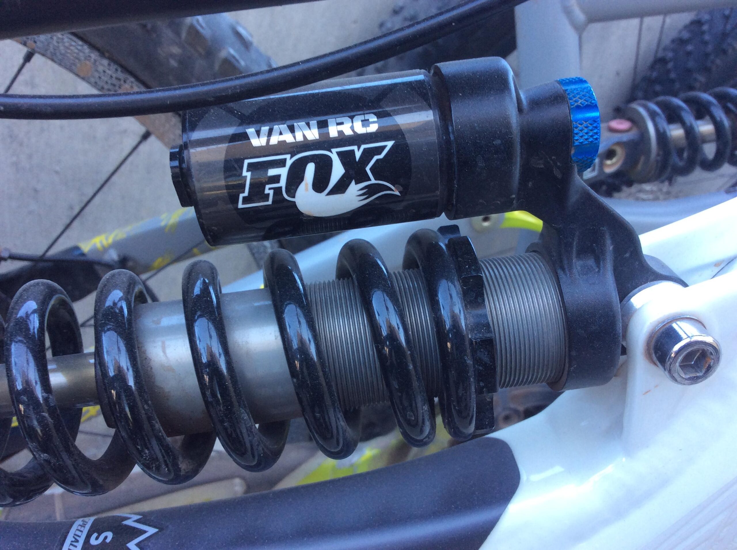 Close-up image of a shock absorber and coil spring assembly mounted on a mountain bike frame. The shock is labeled "VAN RC FOX" and features a black and silver design with a blue adjustment knob. The surrounding components include metallic springs and parts of the bike's structure.