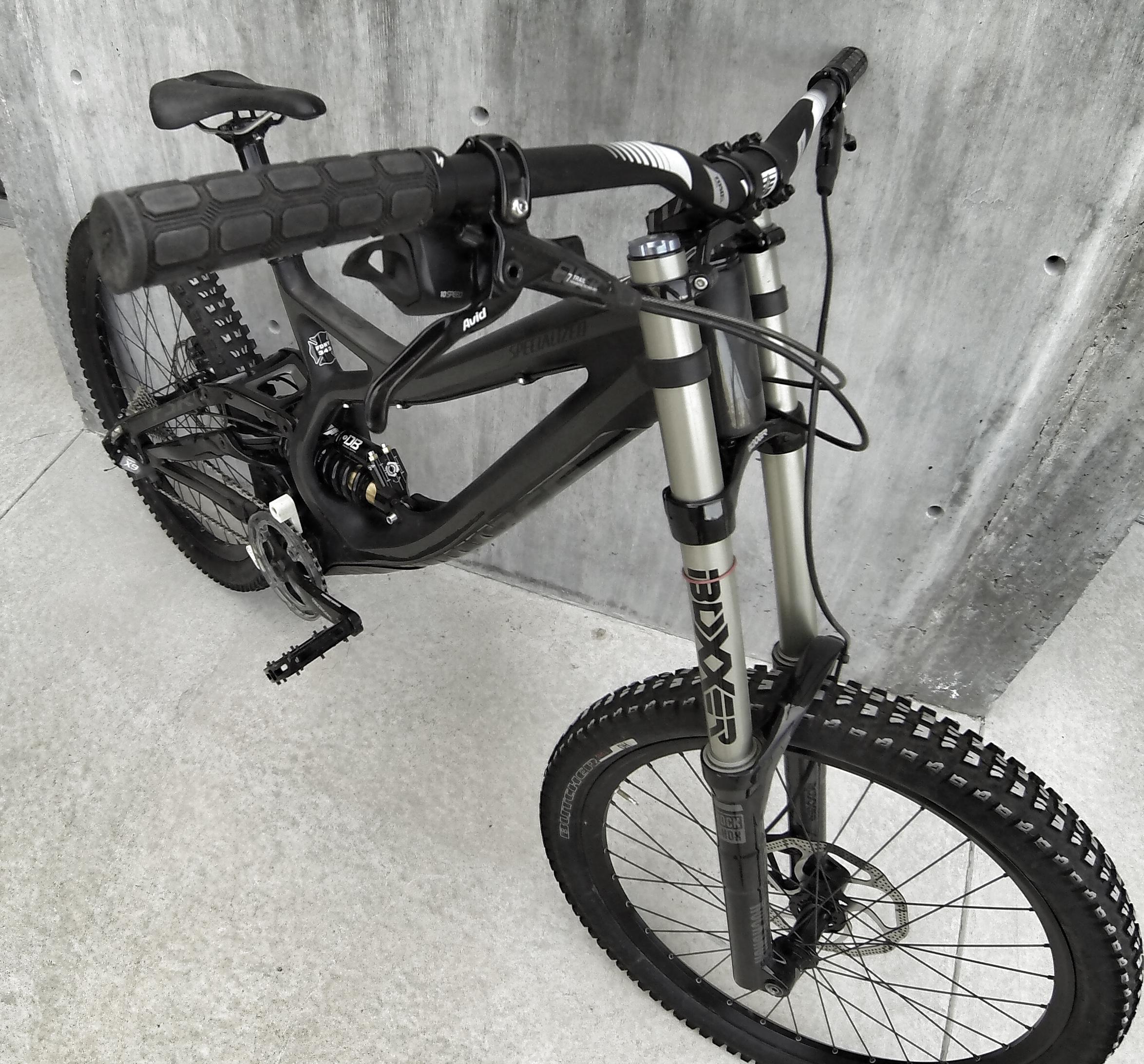 Specialized S-Works Demo 8: A close-up view of a mountain bike resting against a concrete wall. The bike features a dark frame, prominent front suspension forks, and thick, knobby tires, indicating it's designed for off-road cycling. The handlebars are equipped with grips and gear shifters, showcasing its performance-oriented design.