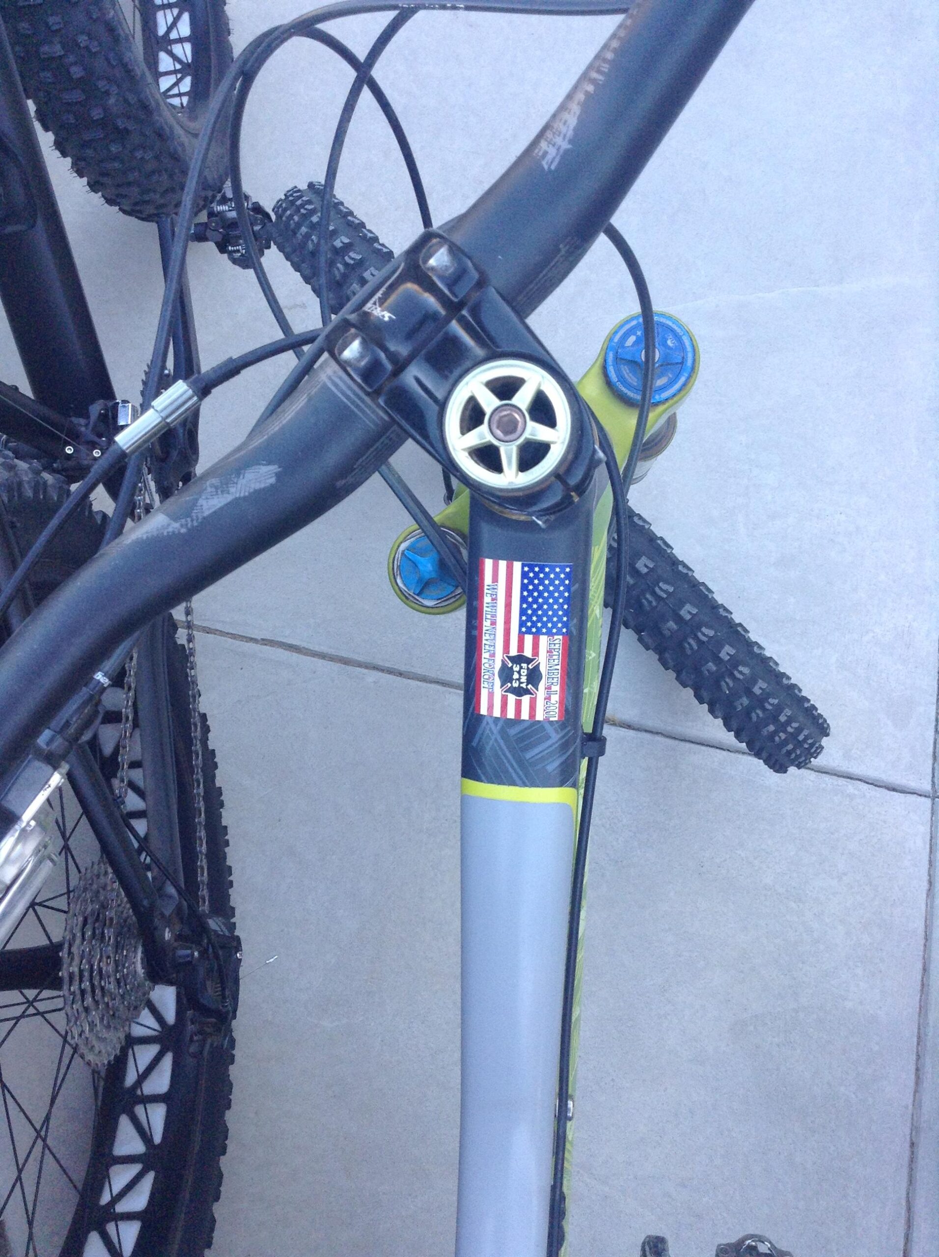 Close-up view of a mountain bike's handlebars and frame, featuring a sticker with the American flag and a skull design. The bike has black handlebars, green accents, and visible cables. In the background, another bike's rear wheel is partially visible against a concrete surface.